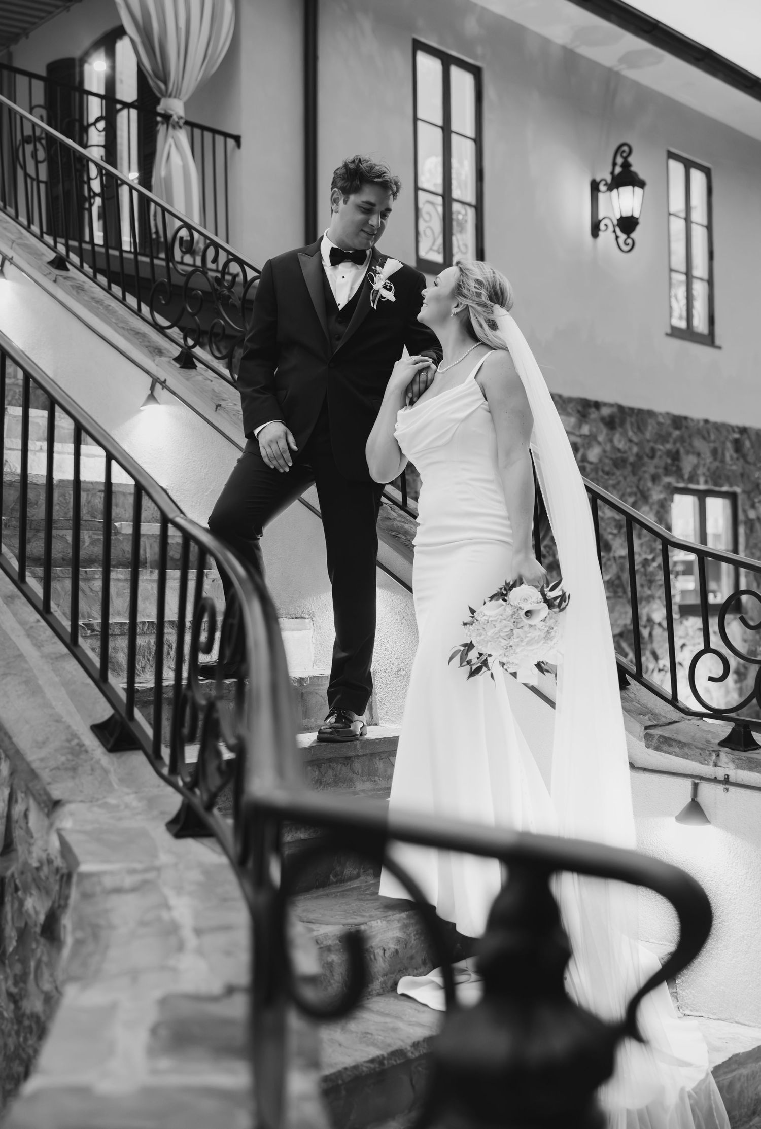Black and white photo of a bride and groom sharing a loving glance on an ornate wrought iron staircase at their wedding venue.