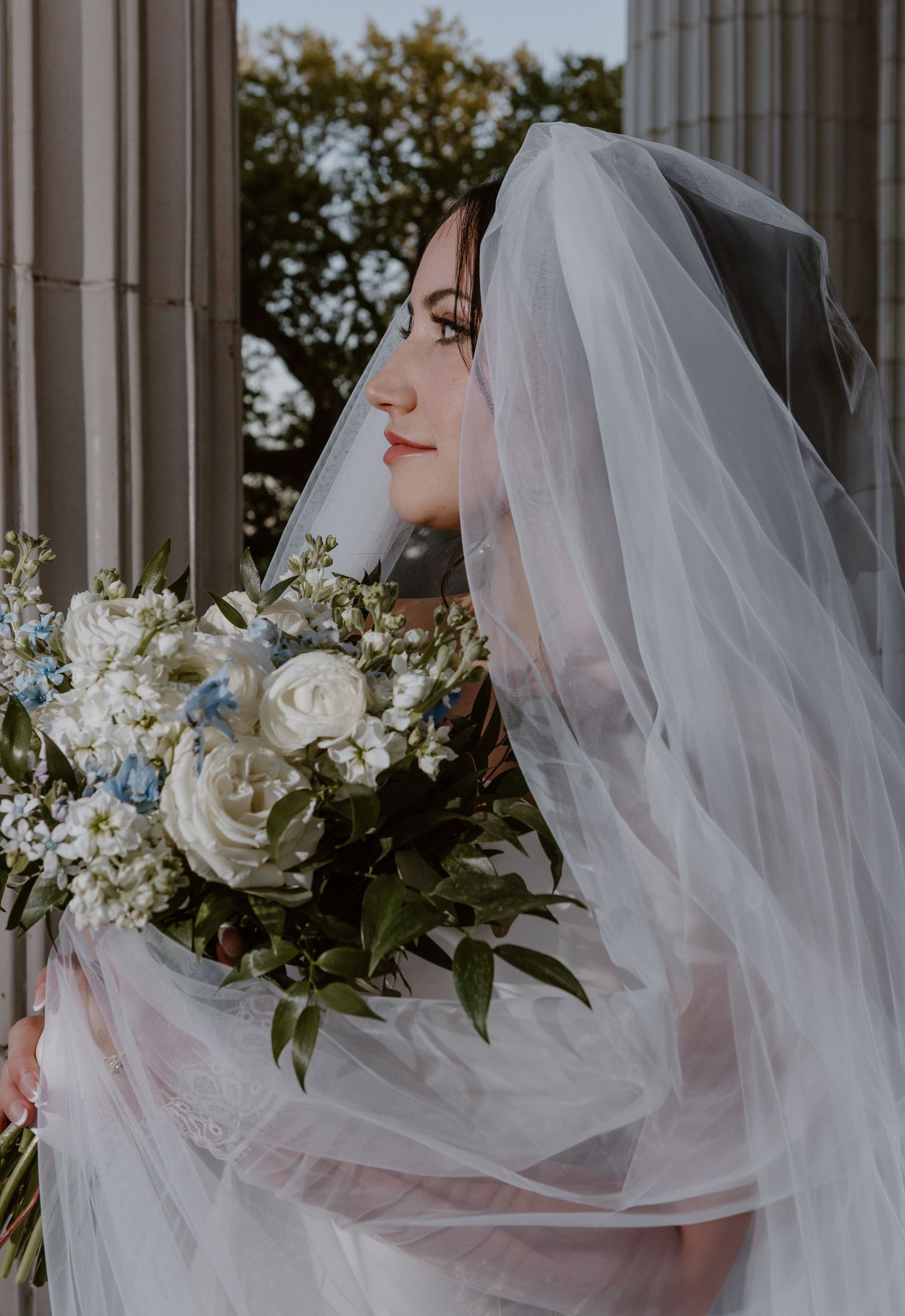 A bride in a flowing white veil holds an elegant white rose and greenery bouquet while smiling serenely against stone columns.