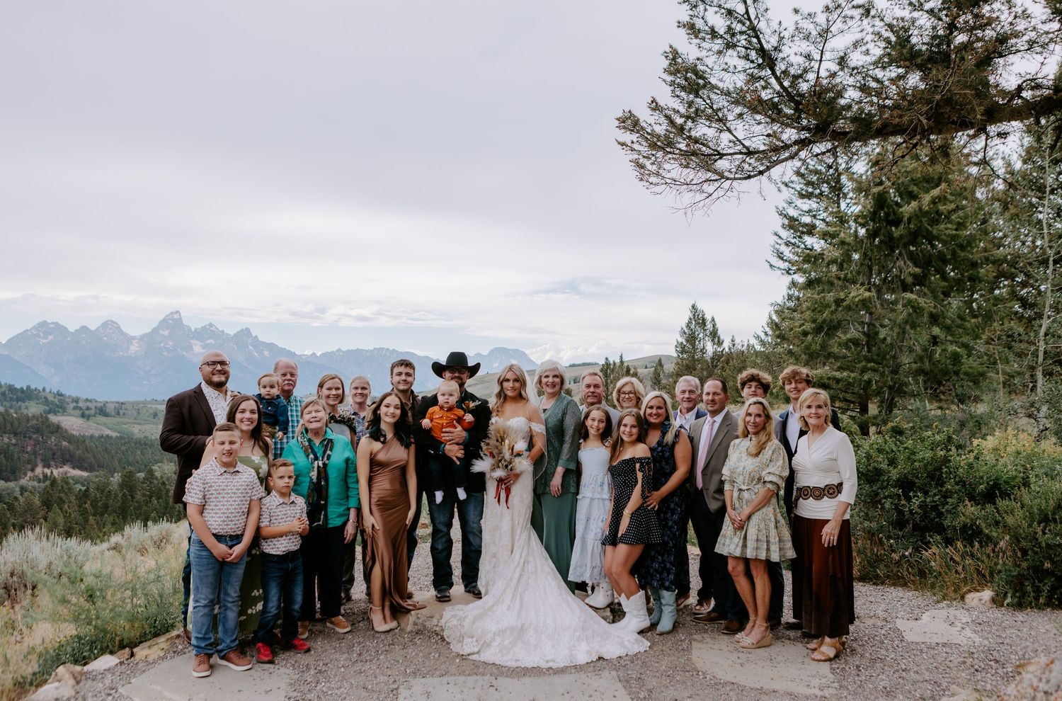 Wedding party poses outdoors with Grand Teton mountains backdrop, bride in white gown, groom in cowboy hat, guests in formal attire.