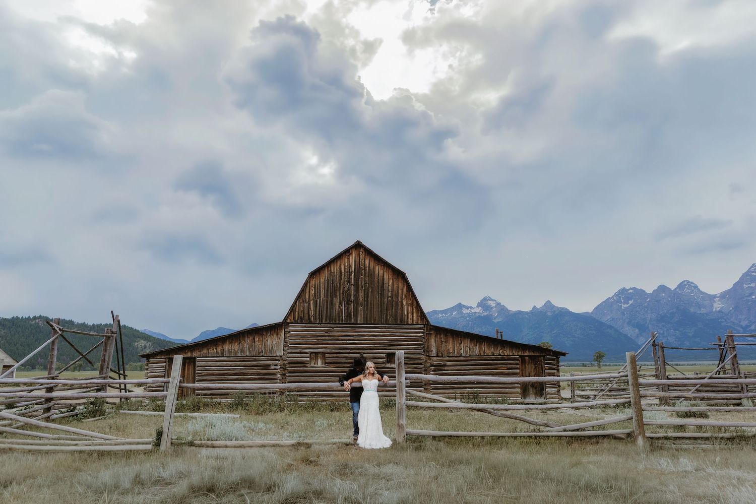 Bride and groom embrace before a rustic barn with Grand Teton mountains under dramatic cloudy skies.