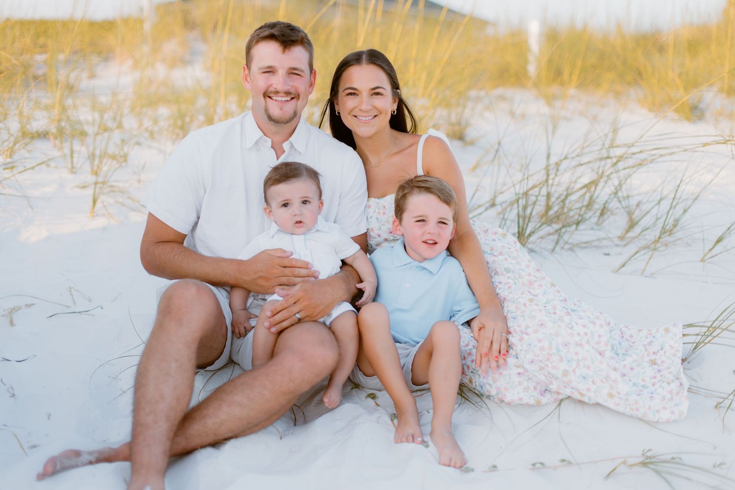 A family of four wearing light colored clothing sits together on white sand dunes at the beach.