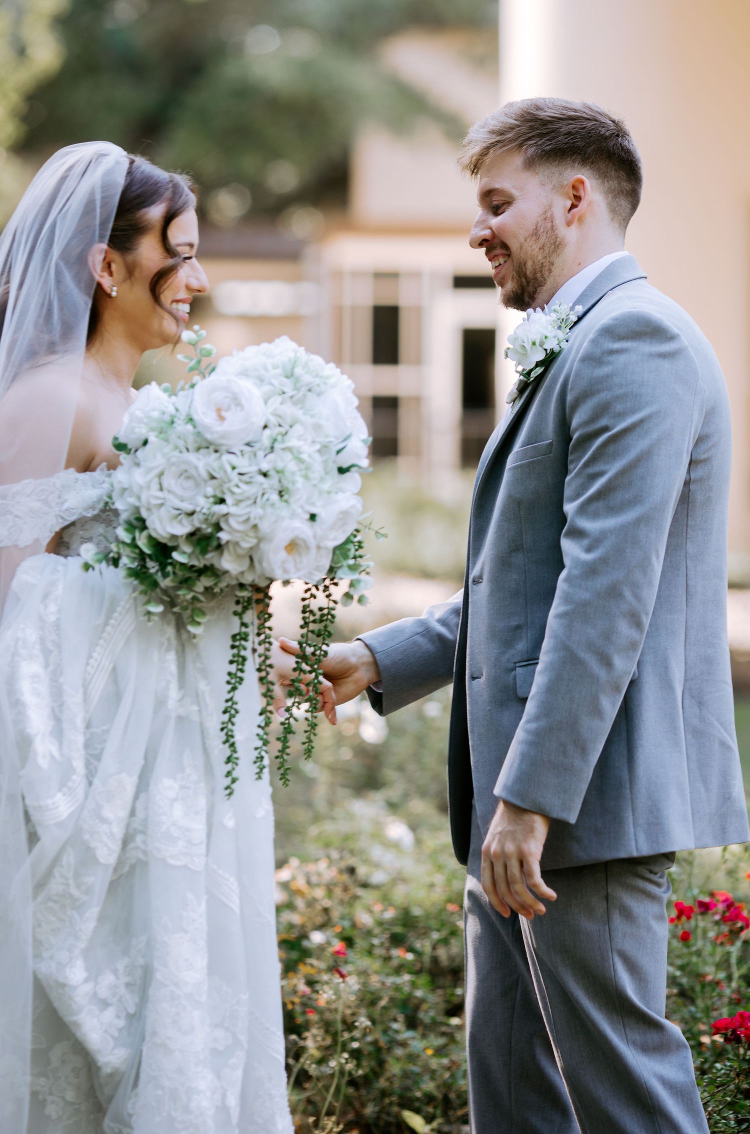 Bride in lace gown holding white rose bouquet smiles at groom in gray suit during outdoor wedding portrait.