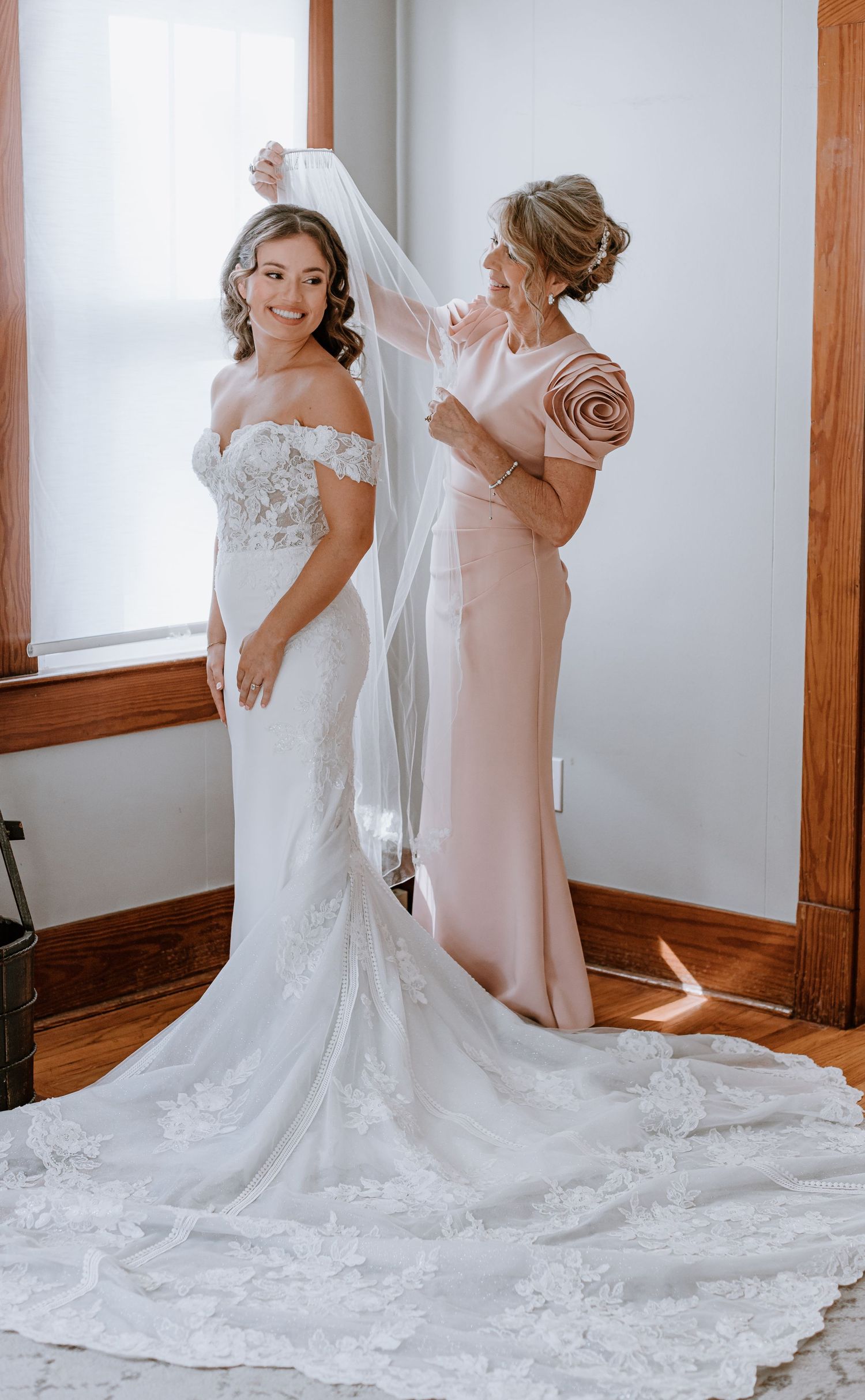 Bride in lace off-shoulder gown with cathedral veil, assisted by woman in blush dress near window.