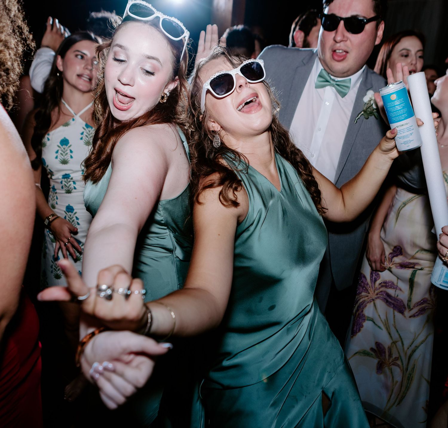 Two young women in green satin dresses dancing and laughing together at a fun wedding reception.