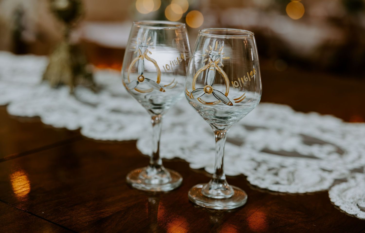 Two decorated wedding wine glasses with gold accents sit on a white lace table runner atop a polished wooden table.