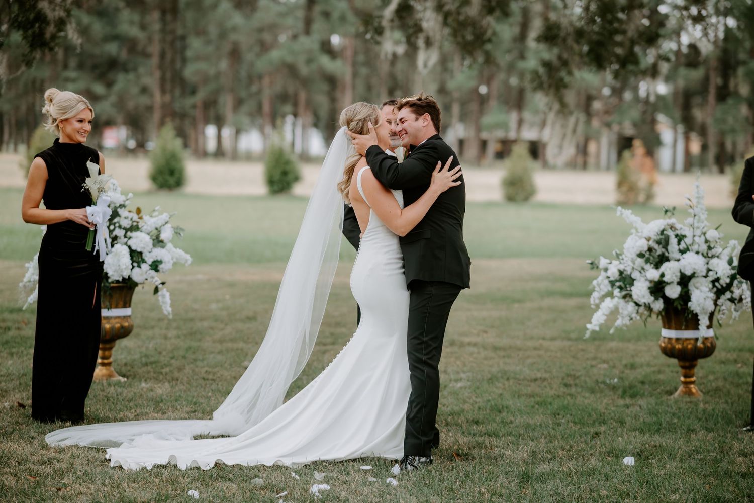 Bride in a long white gown embraces her groom in a black suit during an outdoor garden wedding ceremony with white floral arrangements.