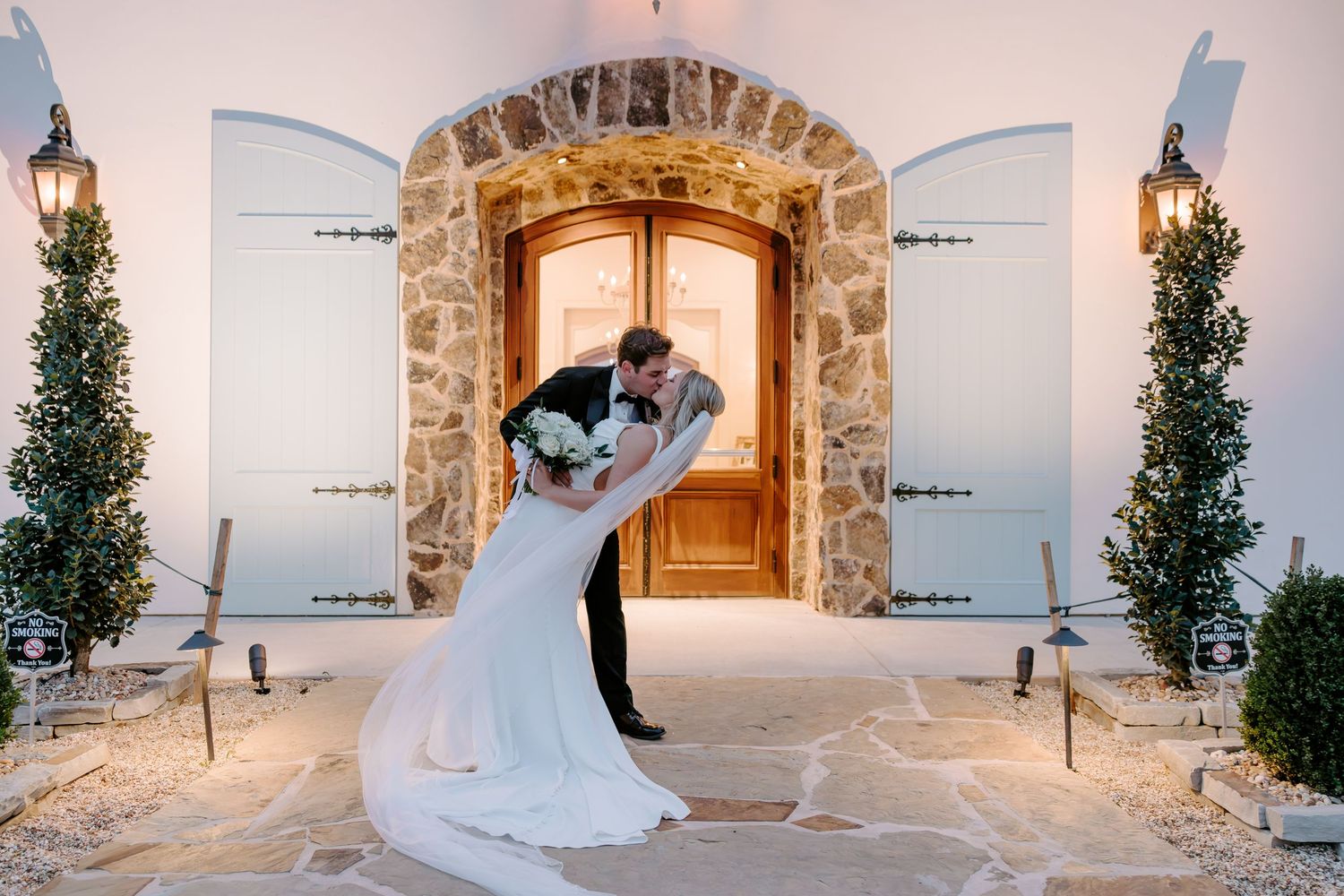 Groom dips and kisses his bride in a white gown at dusk before a rustic stone archway with wooden doors at a wedding venue.