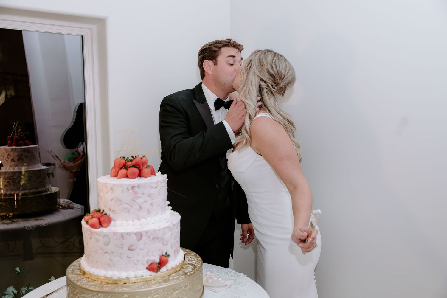 Bride in a white gown and groom in a black tuxedo kiss beside a pink two-tier wedding cake topped with fresh strawberries.