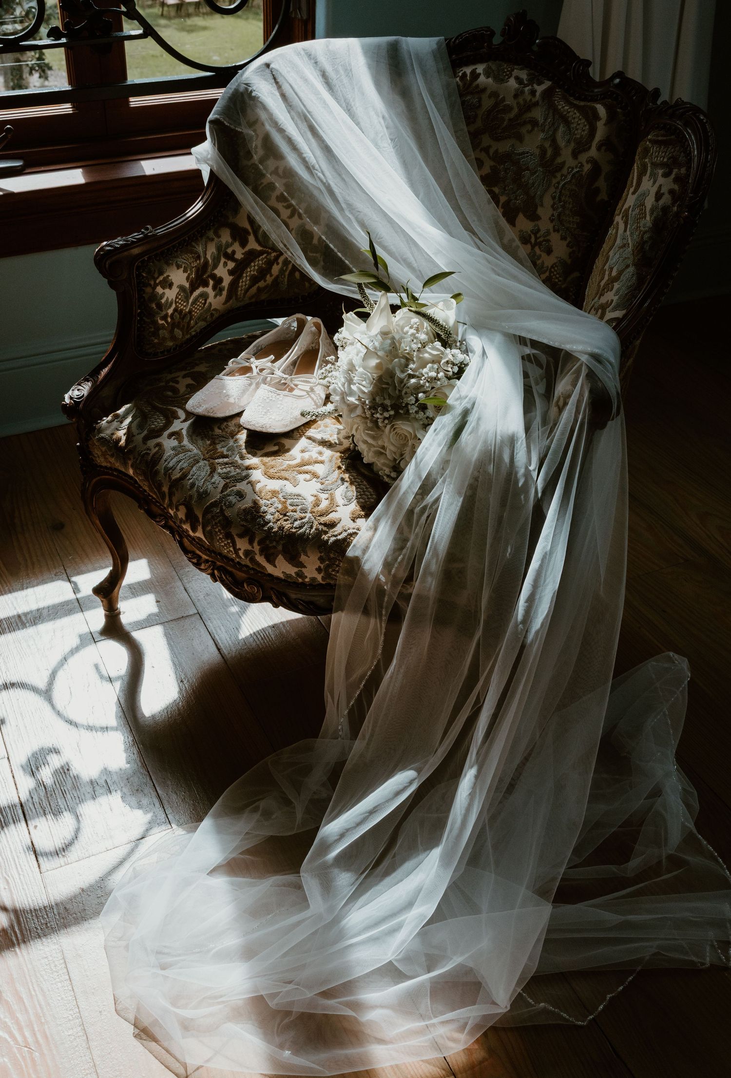 Bridal veil draped over an ornate vintage armchair with ivory ballet flats and a white floral bouquet in soft window light.
