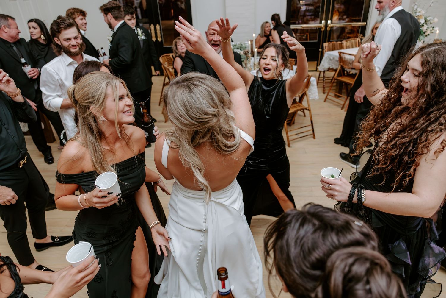 Bride in a white dress dancing with arms raised as smiling wedding guests in black attire cheer and celebrate around her on the dance floor.