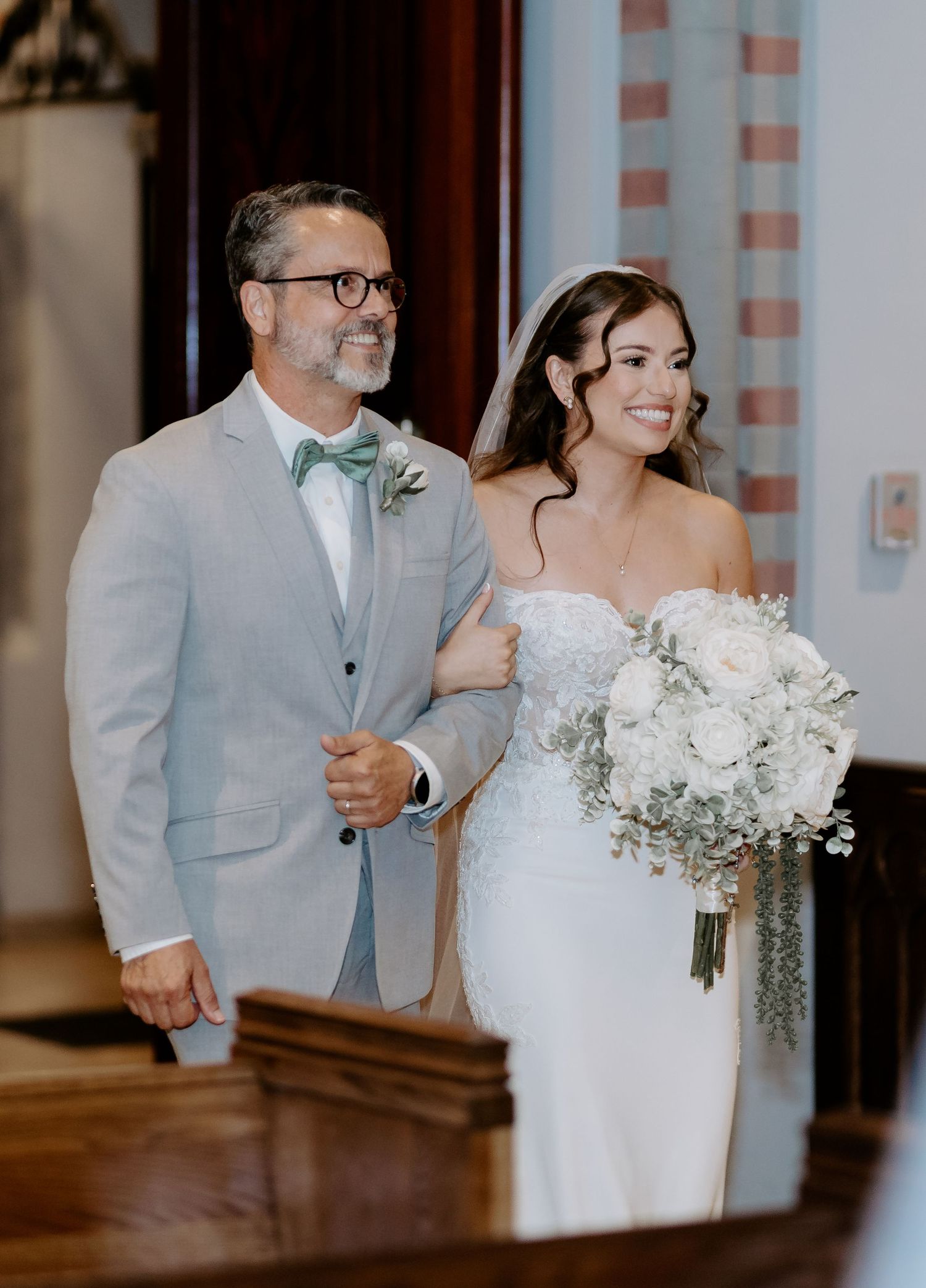 Bride in strapless lace gown holding white bouquet walks down aisle with man in light gray suit and mint bow tie.