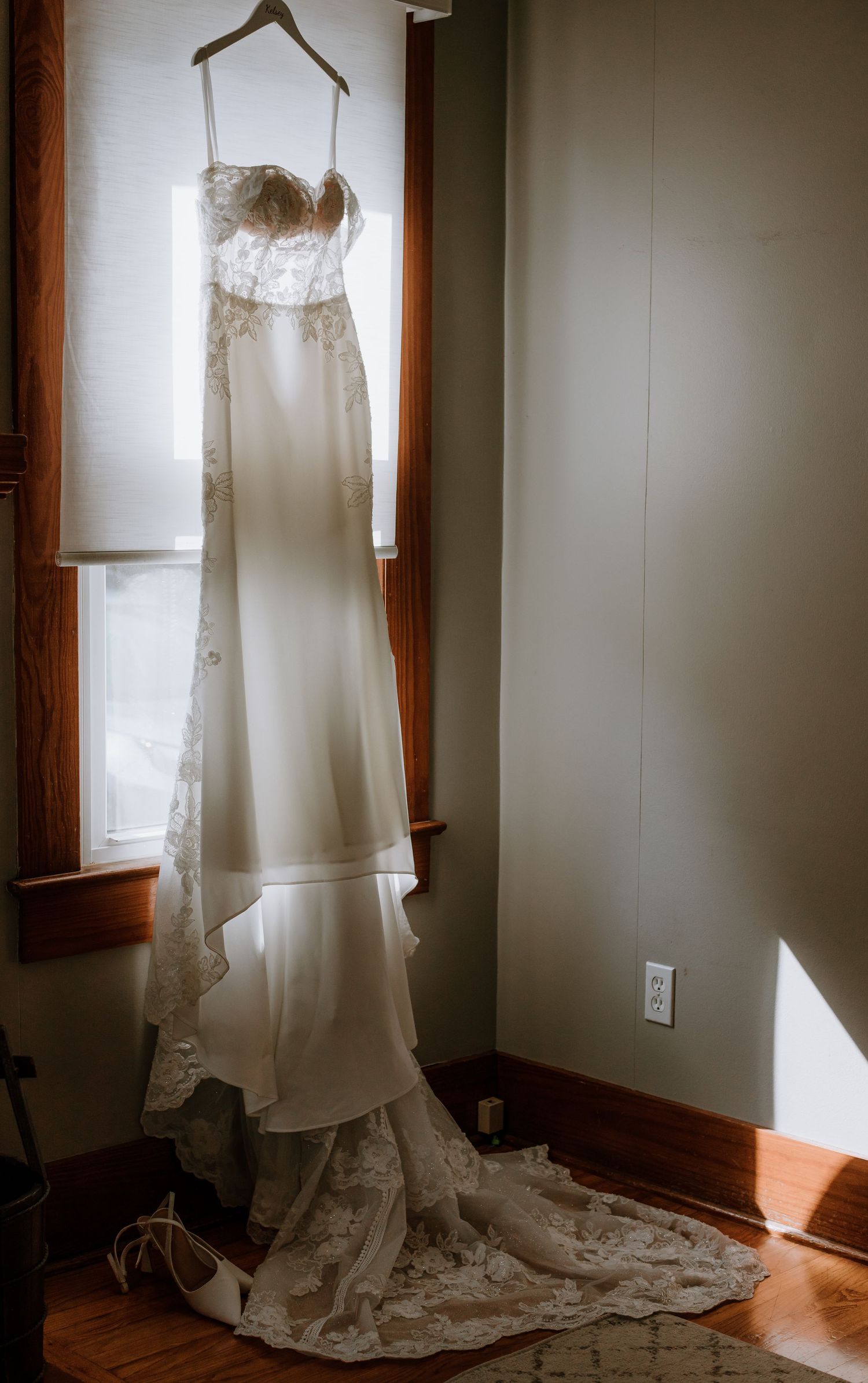 Elegant lace wedding gown hanging by a sunlit window with a flowing train on a wooden floor.