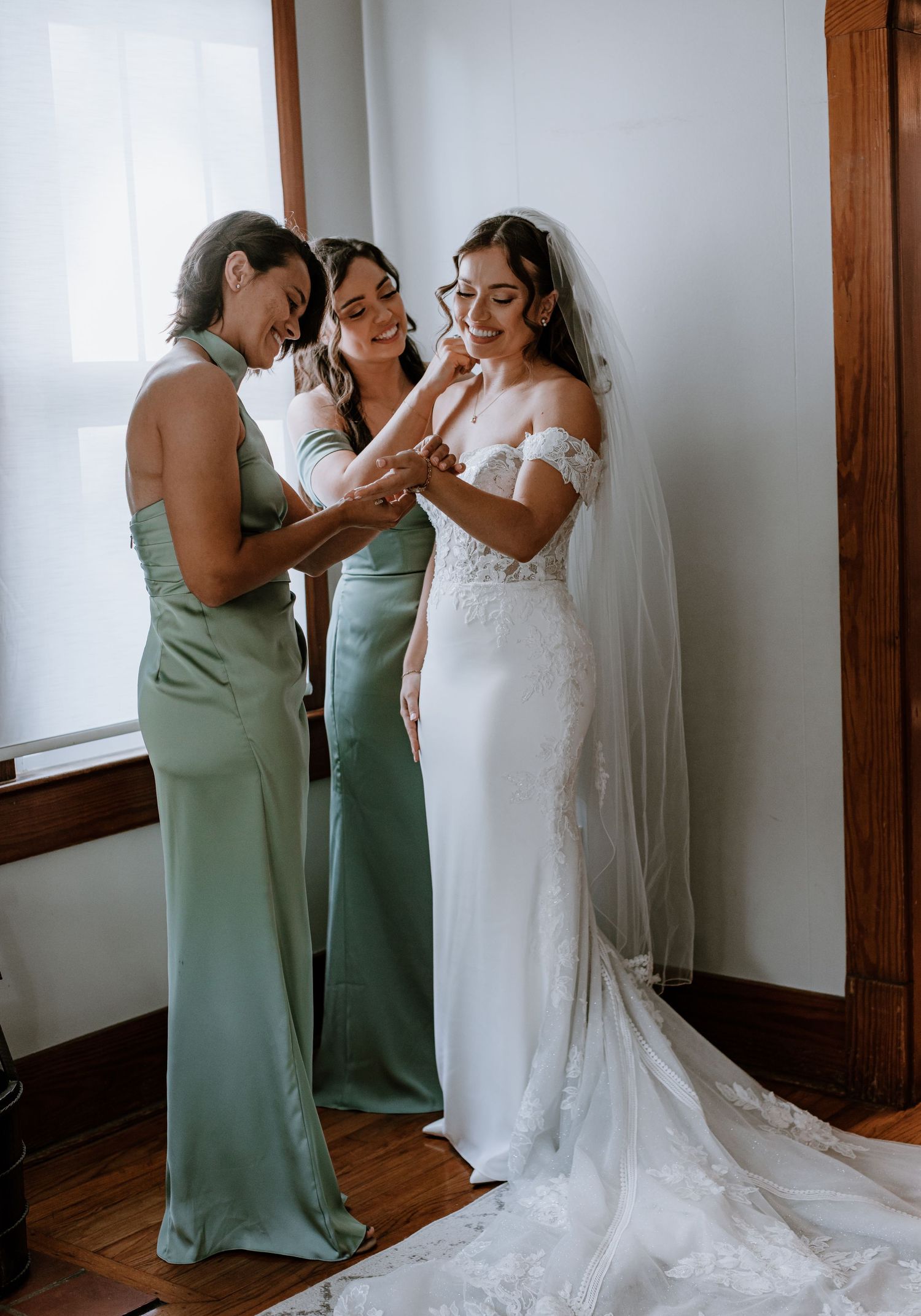 Bride in white lace gown with veil laughs with two bridesmaids in sage green dresses during wedding preparations.