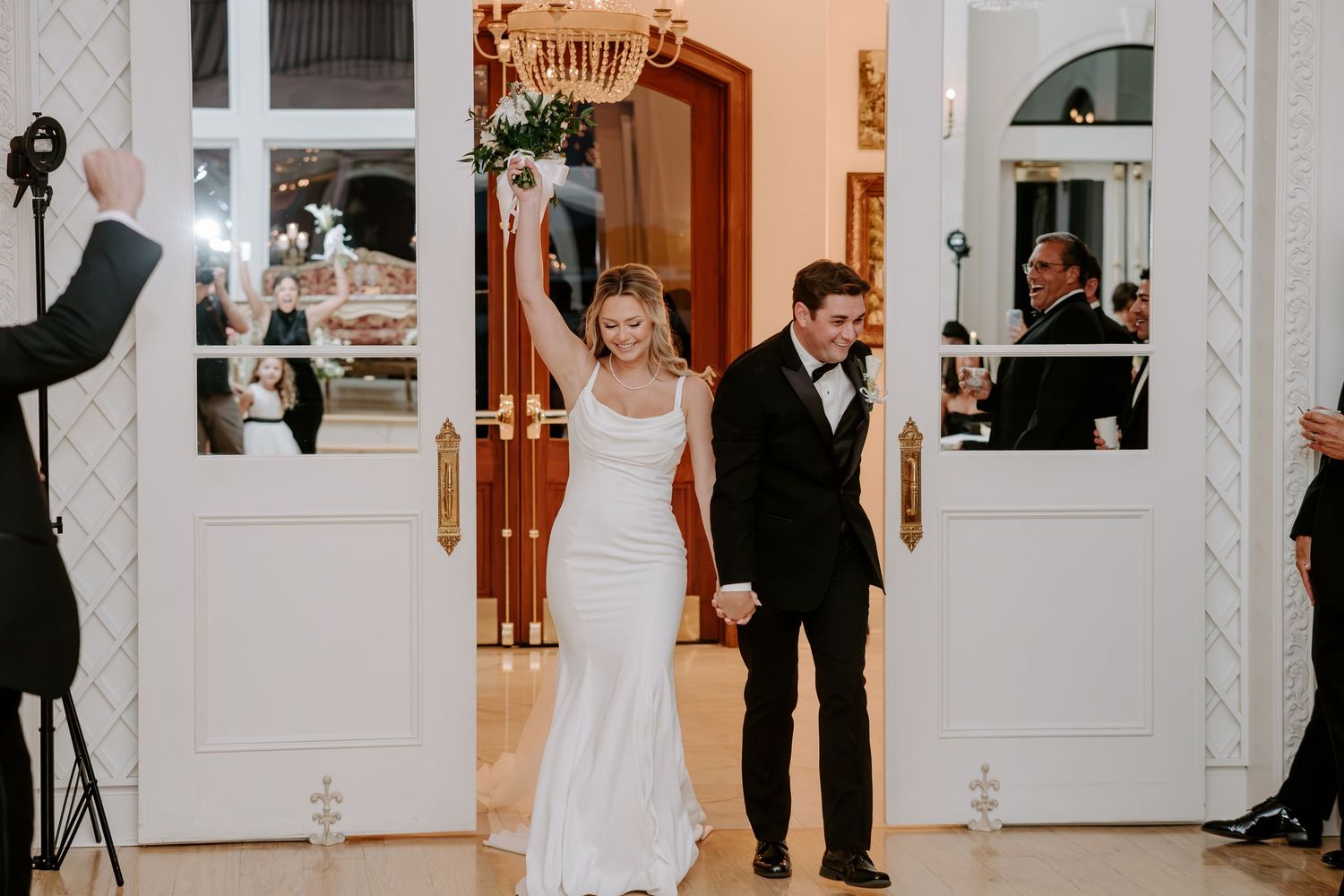 Smiling bride in a white gown and groom in a black tuxedo hold hands and walk into a reception as guests and a photographer look on.
