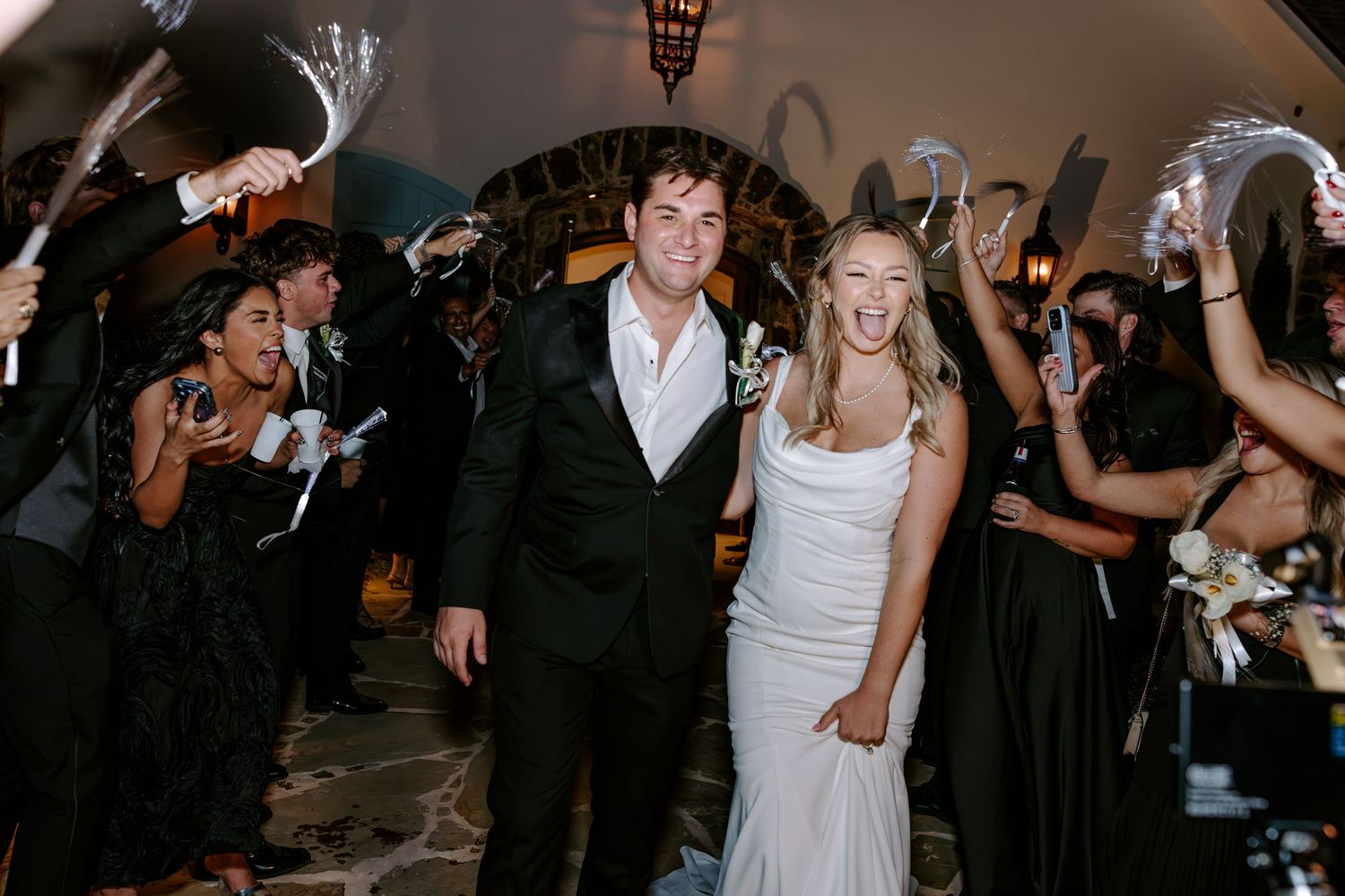 Smiling bride in a white gown and groom in a black suit walk through cheering wedding guests holding sparklers at an evening send-off.