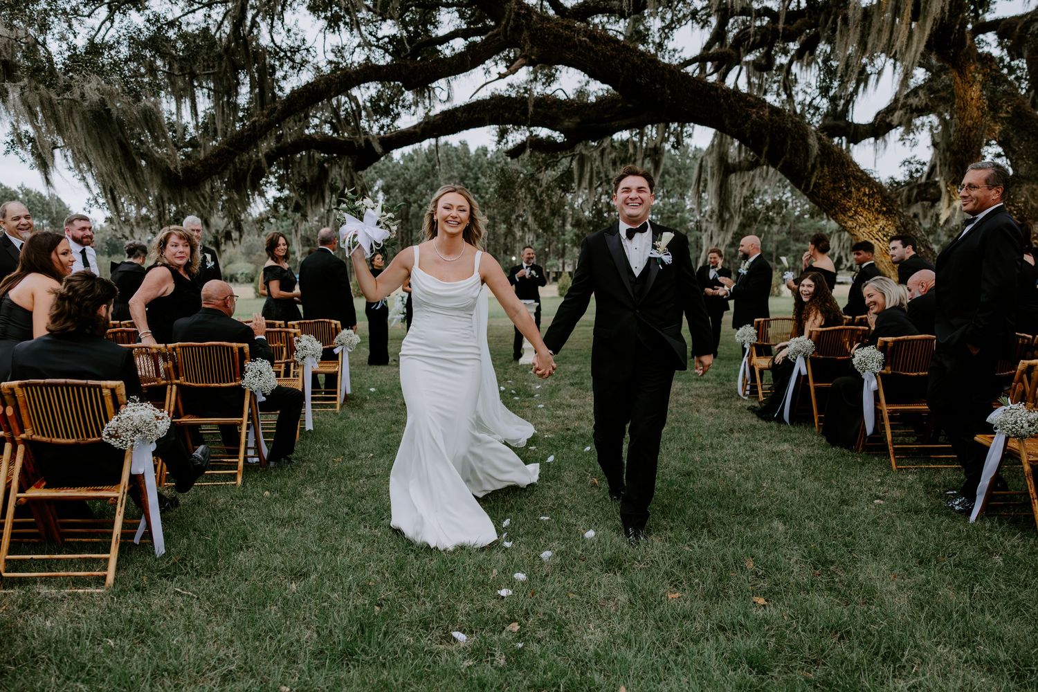 Smiling bride and groom hold hands and walk down the aisle beneath large oak trees as seated wedding guests cheer at an outdoor ceremony.