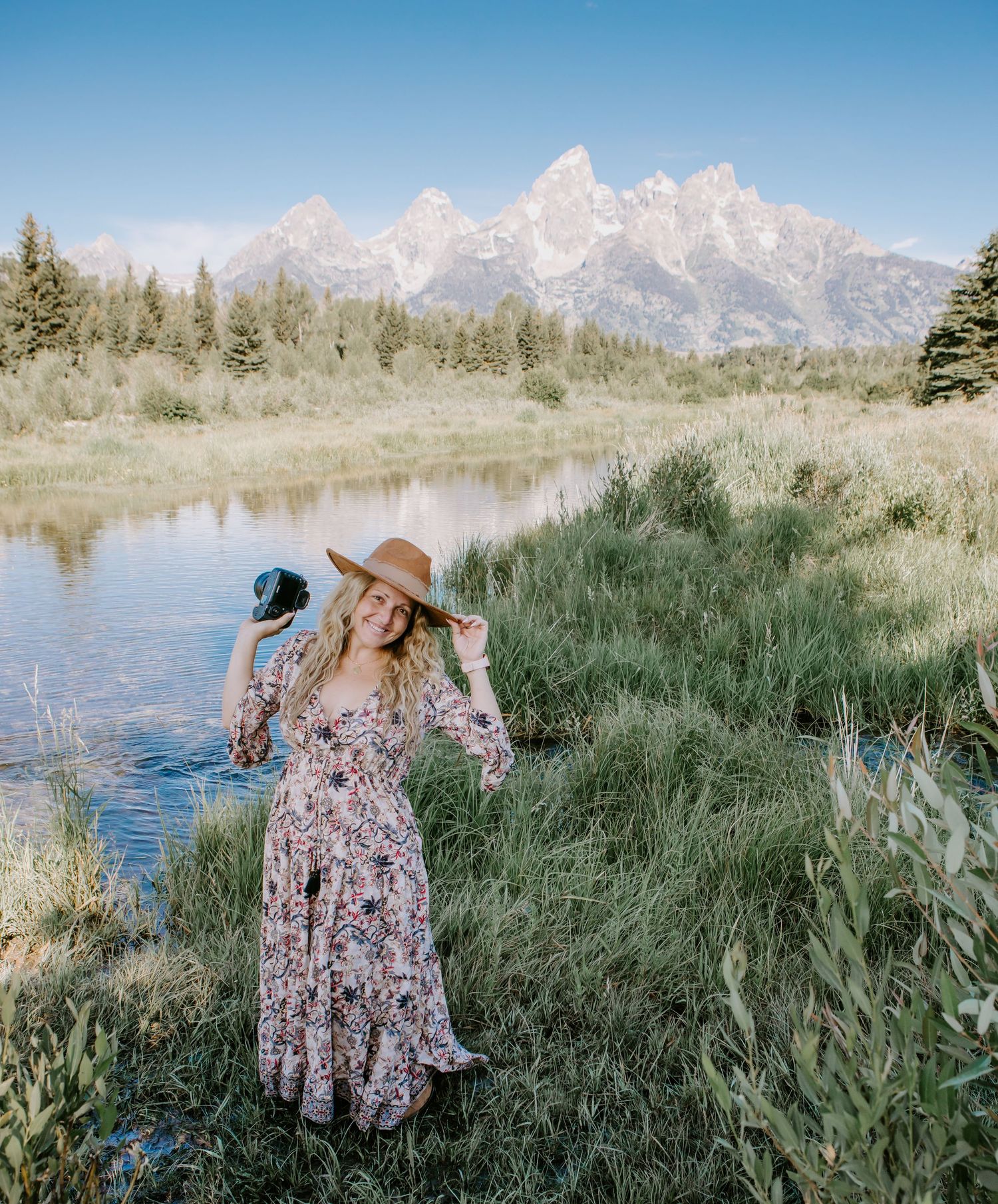 Woman in floral maxi dress and sun hat poses by a river with Grand Teton mountains in the background.