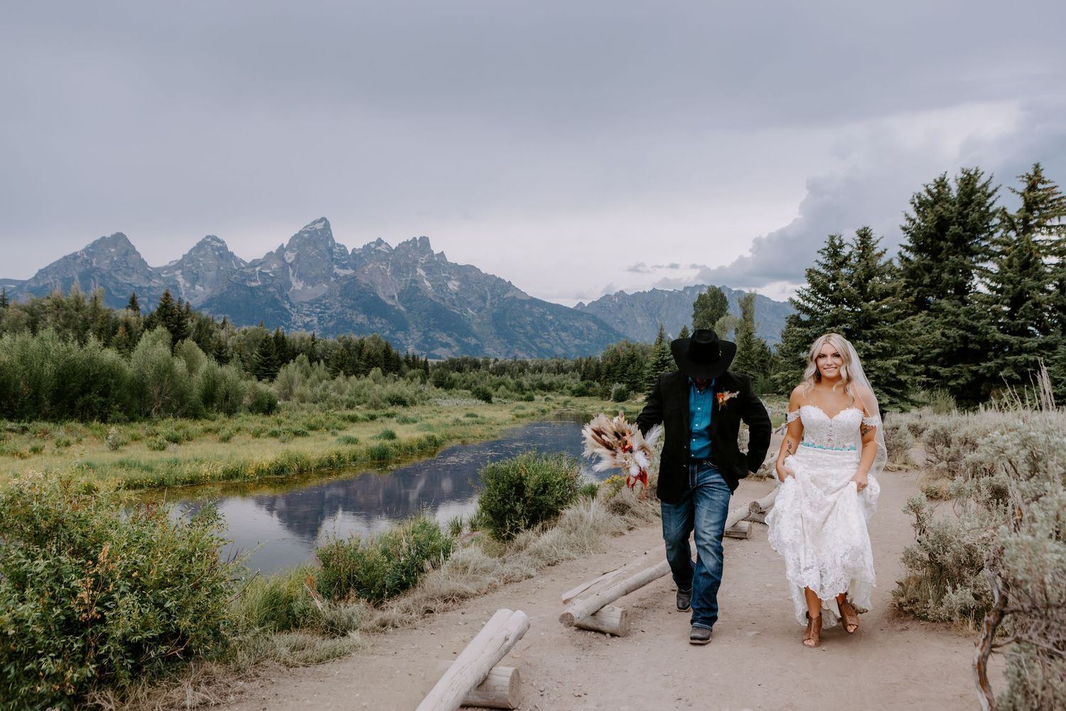 Bride and groom walk hand-in-hand on a scenic mountain path near a river in Grand Teton National Park.