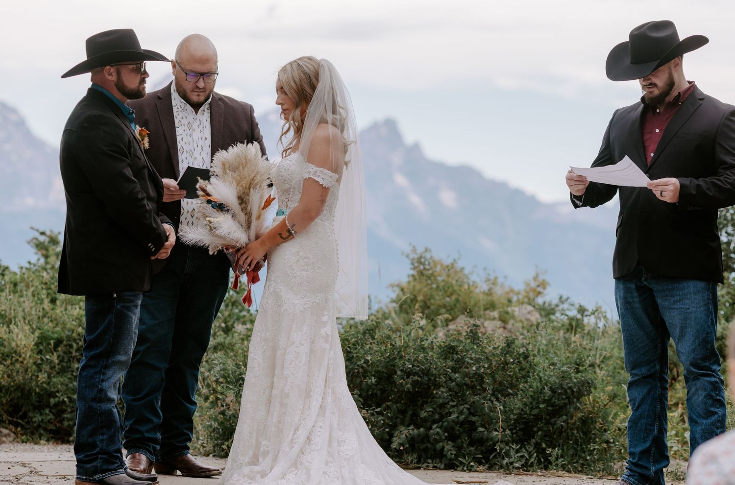 Outdoor mountain wedding ceremony with bride, groom, officiant, and guest in cowboy hats, with a white dog.