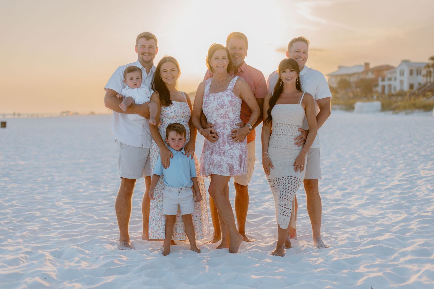 A group poses together on a sandy beach at sunset with coastal homes visible in the distance.