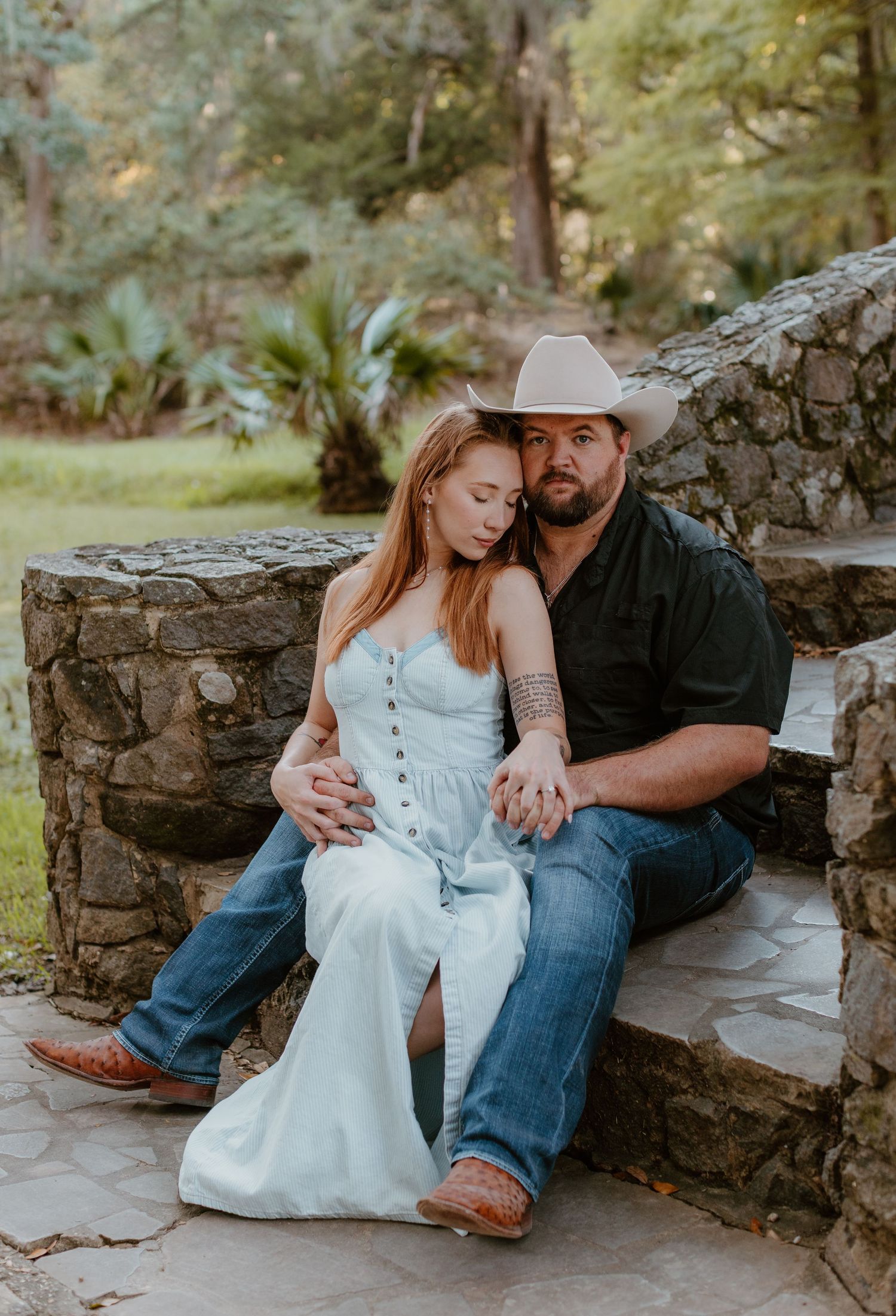 A couple sits together on rustic stone steps in an outdoor setting wearing casual attire during an engagement photoshoot.