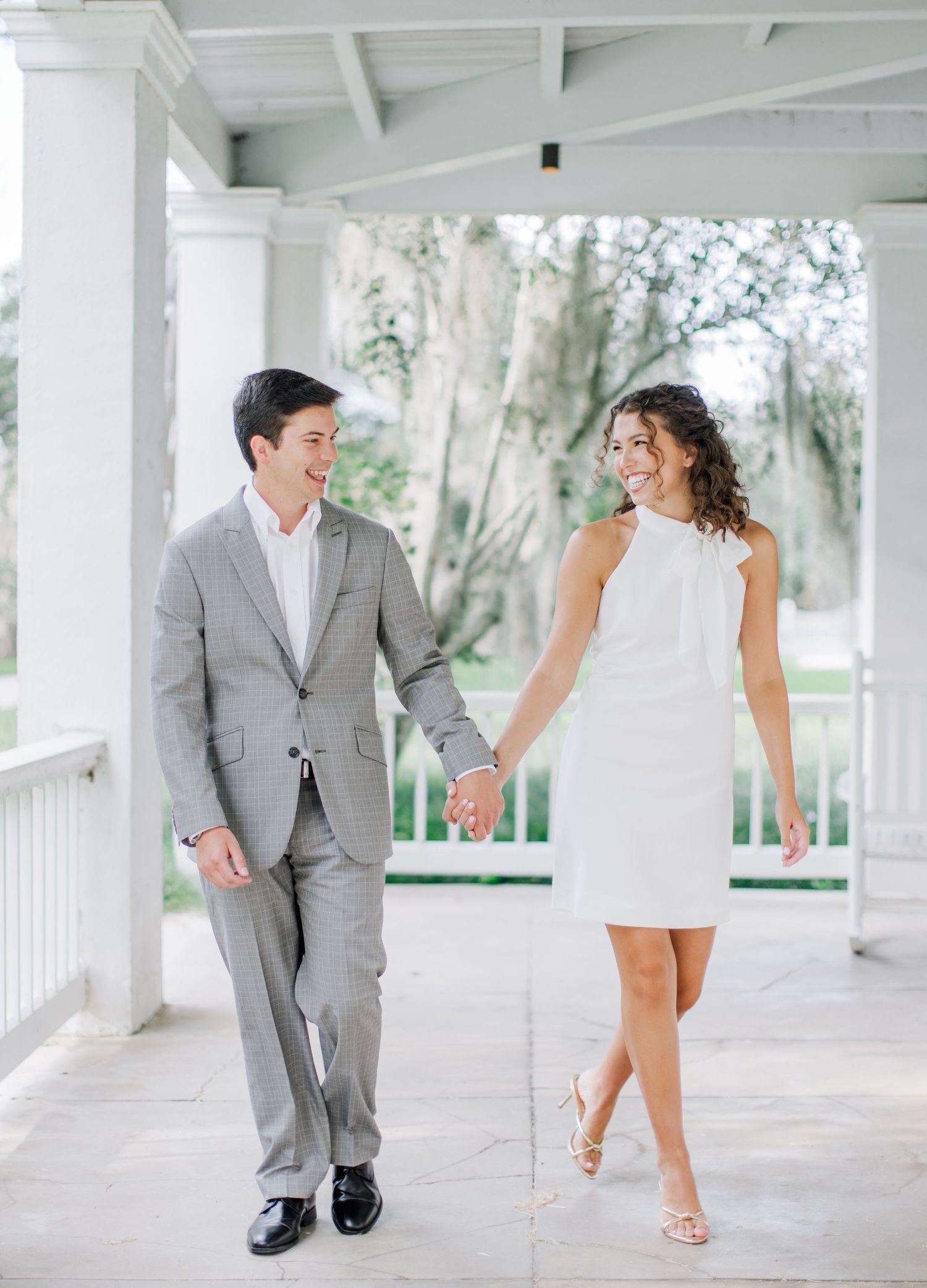 Playful couple in wedding attire share romantic moments under a white columned porch with Spanish moss in background.