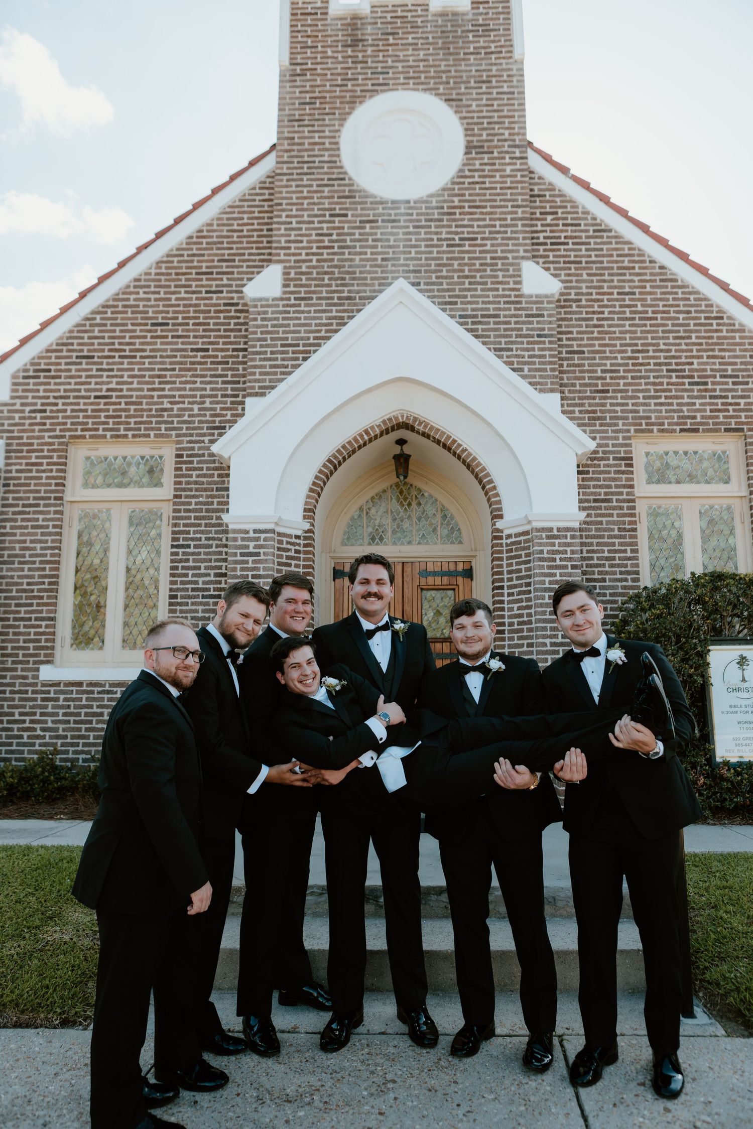 Groomsmen in black tuxedos lifting the groom outside a brick church on his wedding day.