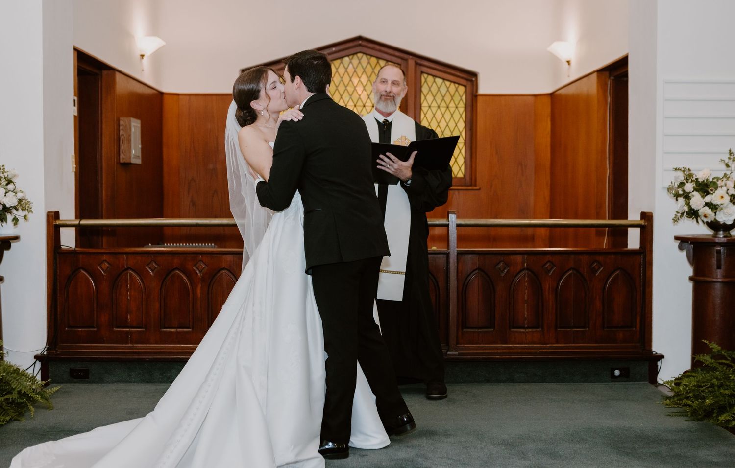 Bride and groom share first kiss at church altar with officiant holding book during elegant wedding ceremony.