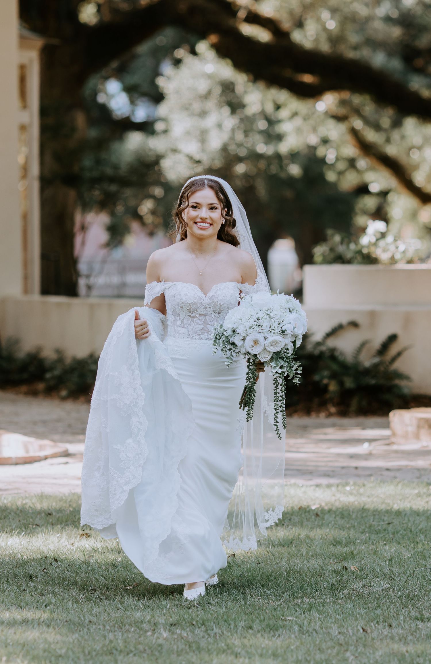 Smiling bride in off-shoulder lace wedding gown holding white bouquet walking outdoors on green lawn.