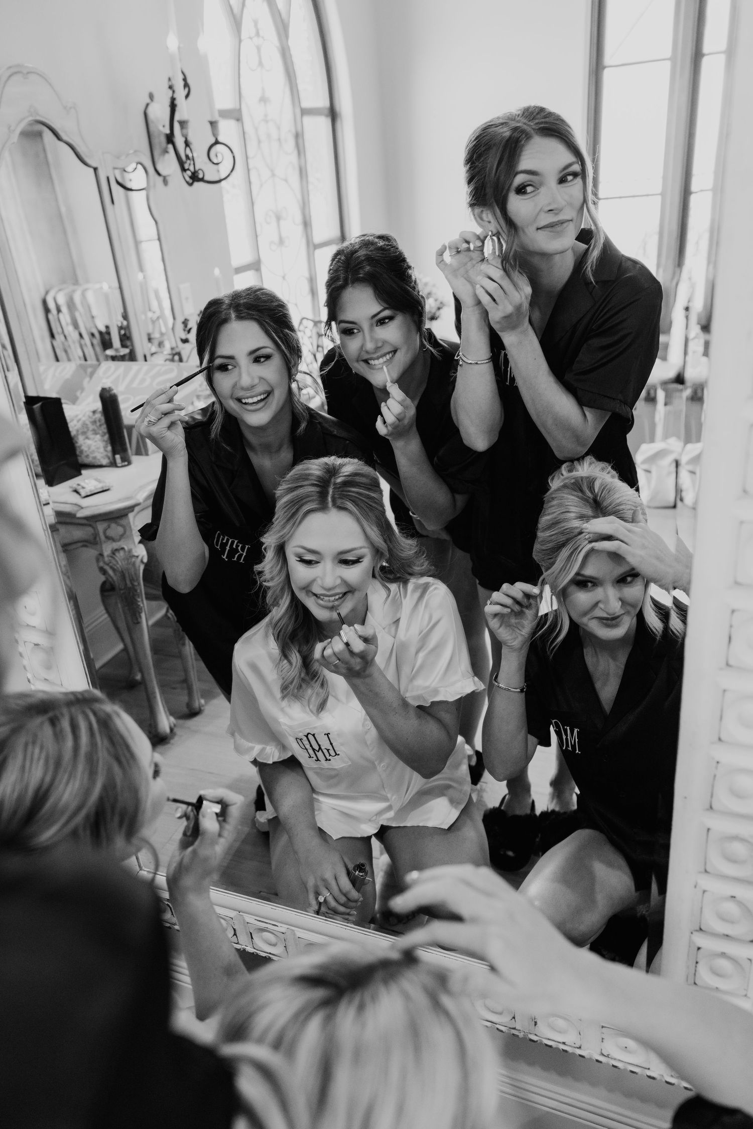 Black and white photo of bridesmaids in matching robes laughing while getting ready together in front of a large mirror.