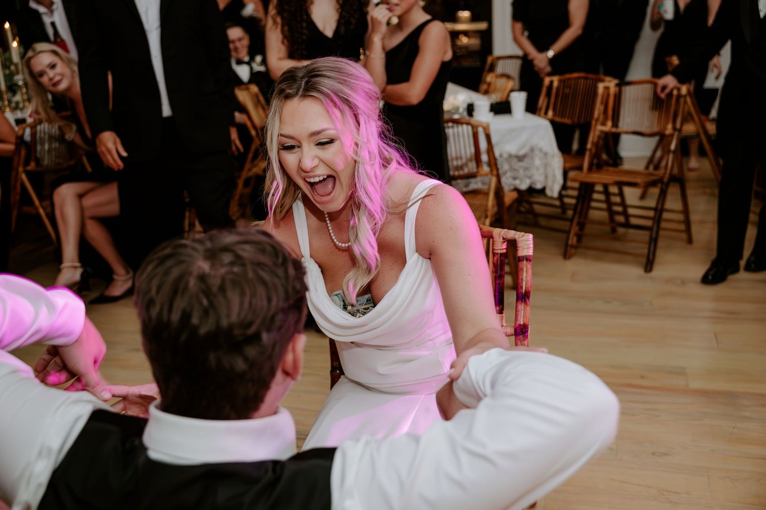 Laughing woman with blonde-pink hair in a white dress is lifted on a chair during a lively wedding reception as guests cheer behind her.