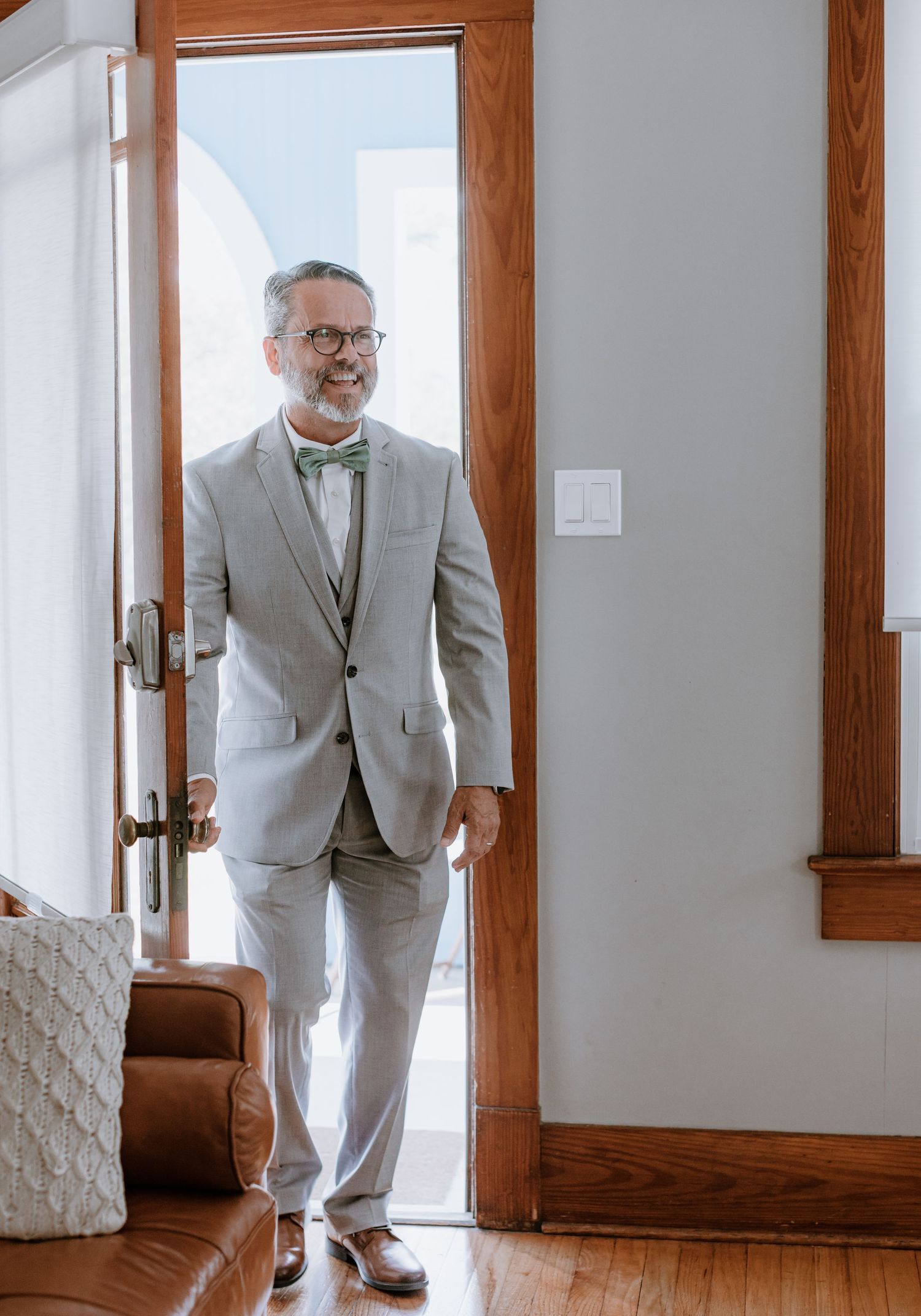 A smiling middle-aged man in a light gray suit and bow tie stands in a wooden doorway on hardwood floors.