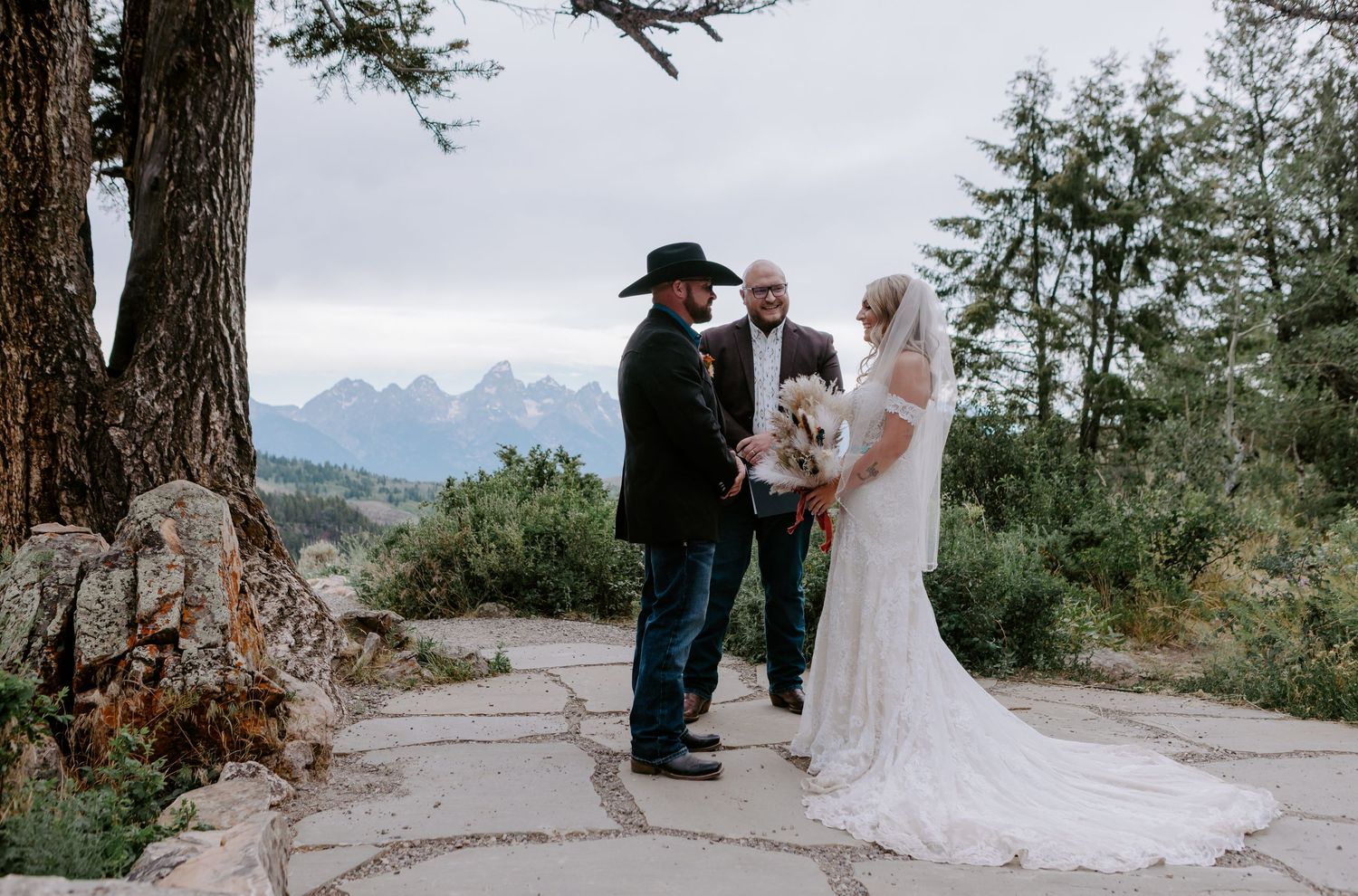 Outdoor wedding ceremony with couple and officiant on stone path, mountain backdrop, bride in white gown, groom in black cowboy hat.