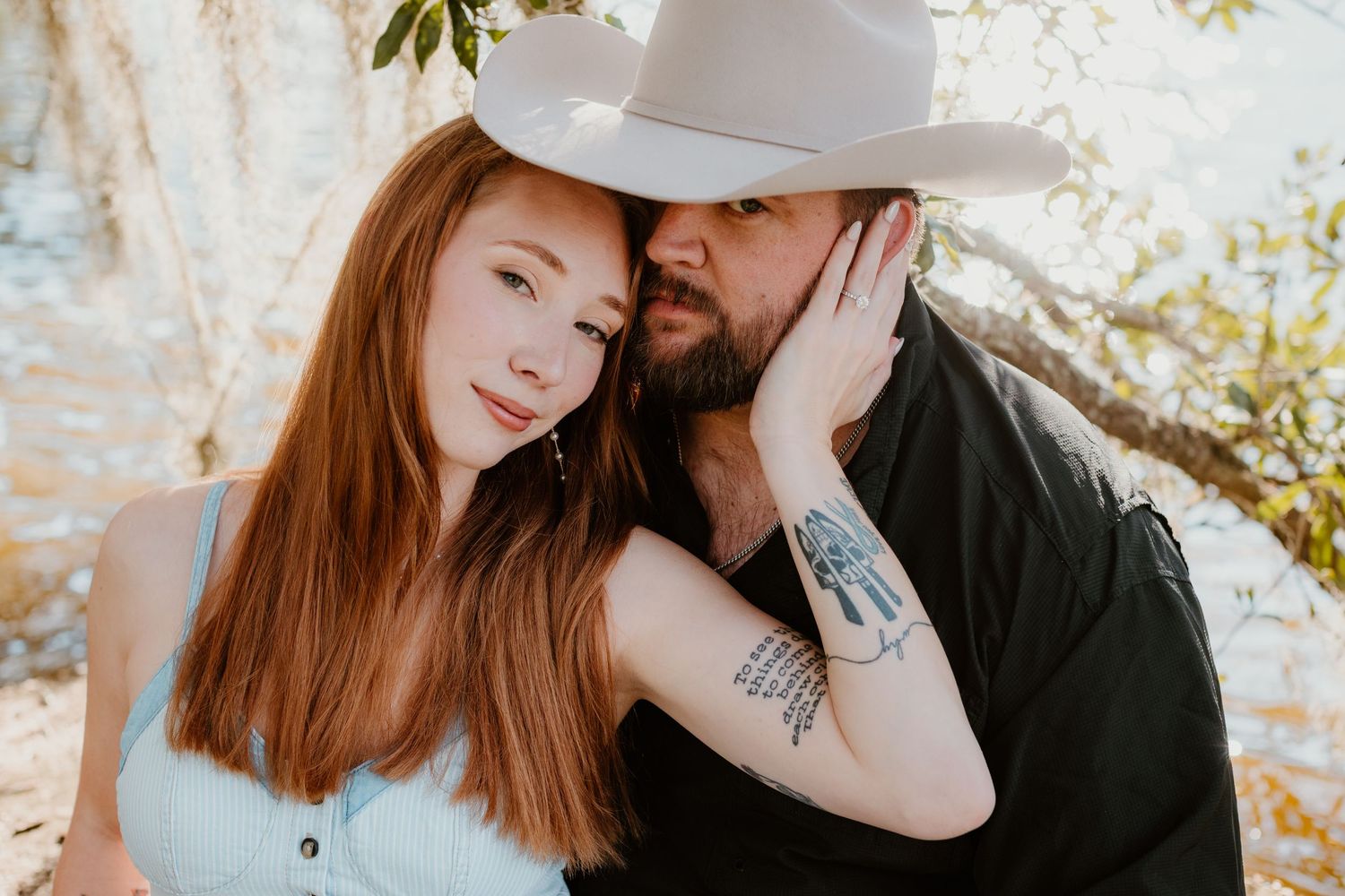 A couple in casual attire embraces outdoors on a sunny day with natural foliage in the background.