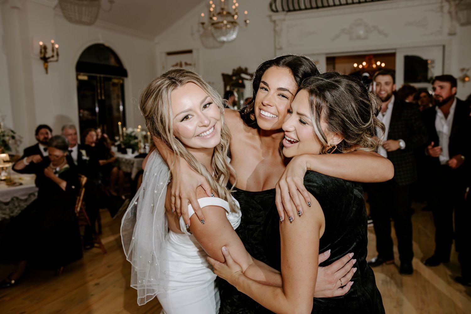 A smiling bride in a white dress embraces two bridesmaids in black dresses during a joyful wedding reception with guests in the background.