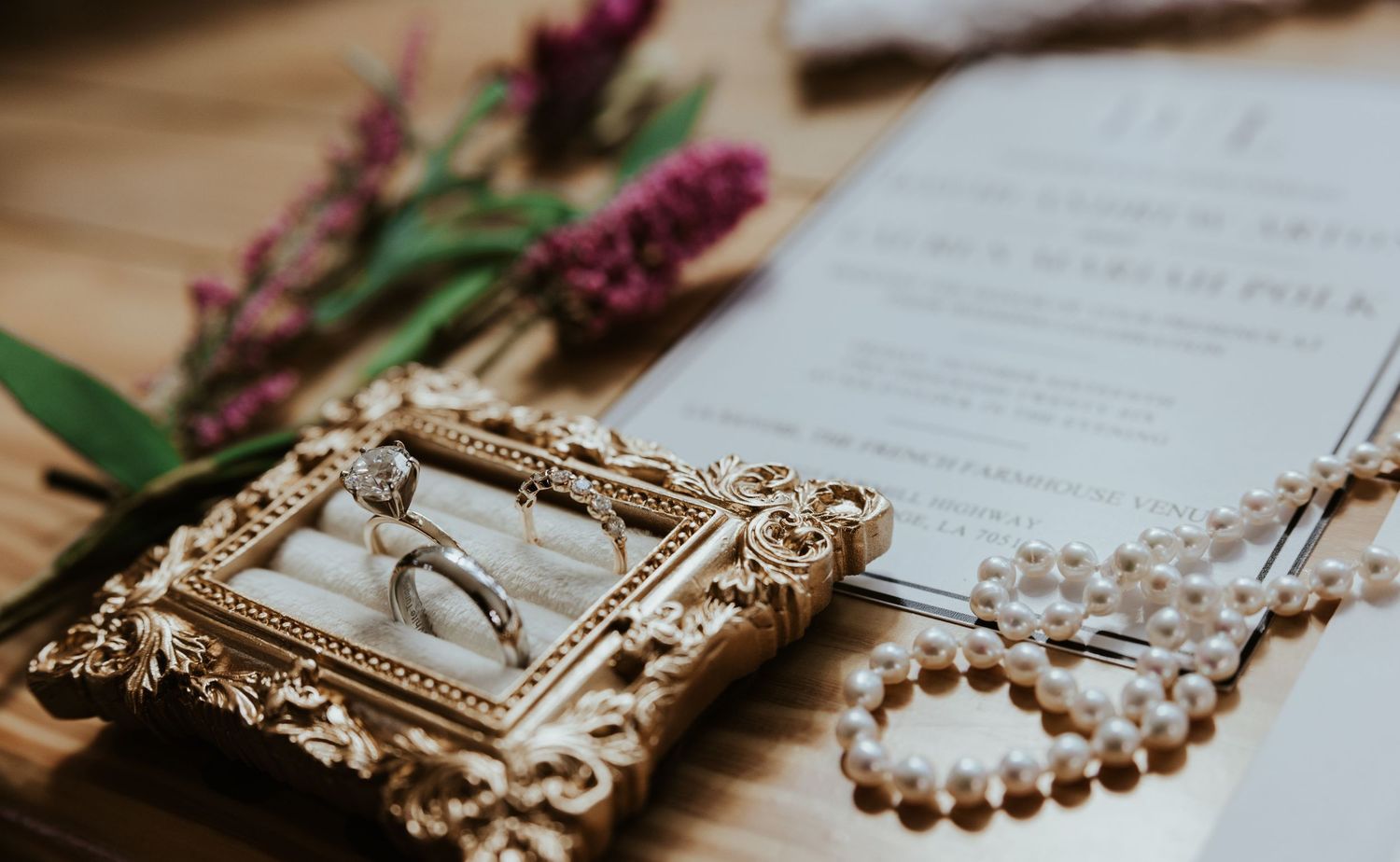Two gold wedding rings in an ornate vintage gold frame ring box beside a pearl bracelet, pink flowers and a wedding invitation.