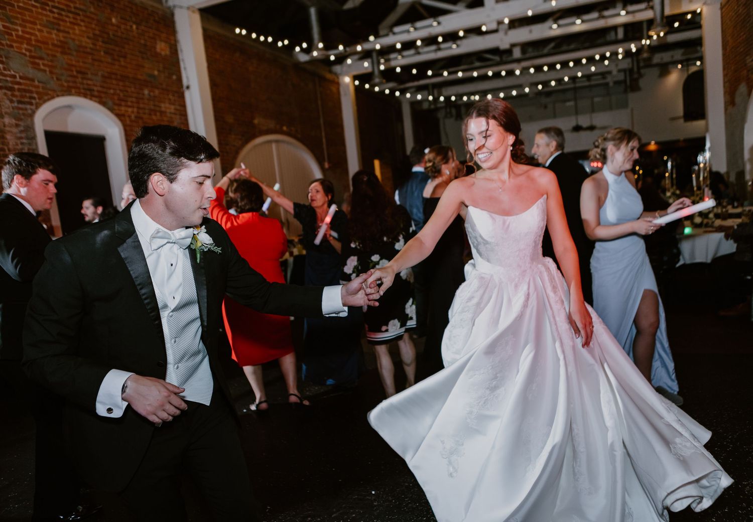 Bride in white gown and groom in tuxedo dance joyfully at their wedding reception under string lights.