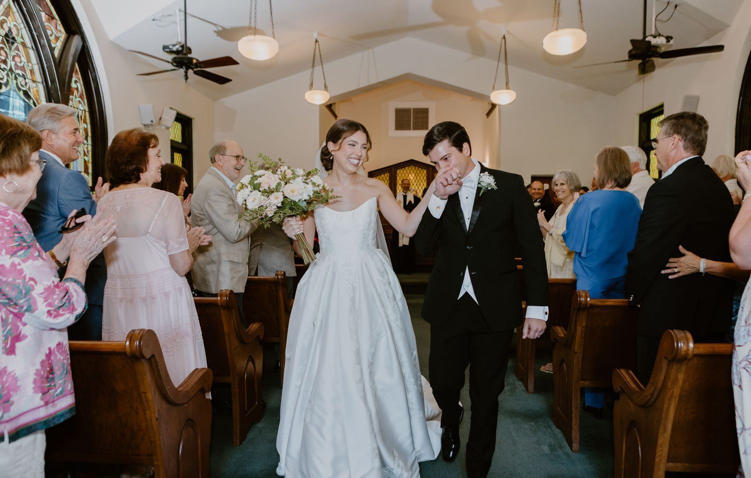 Bride and groom walk down church aisle after wedding ceremony, guests applauding in wooden pews.