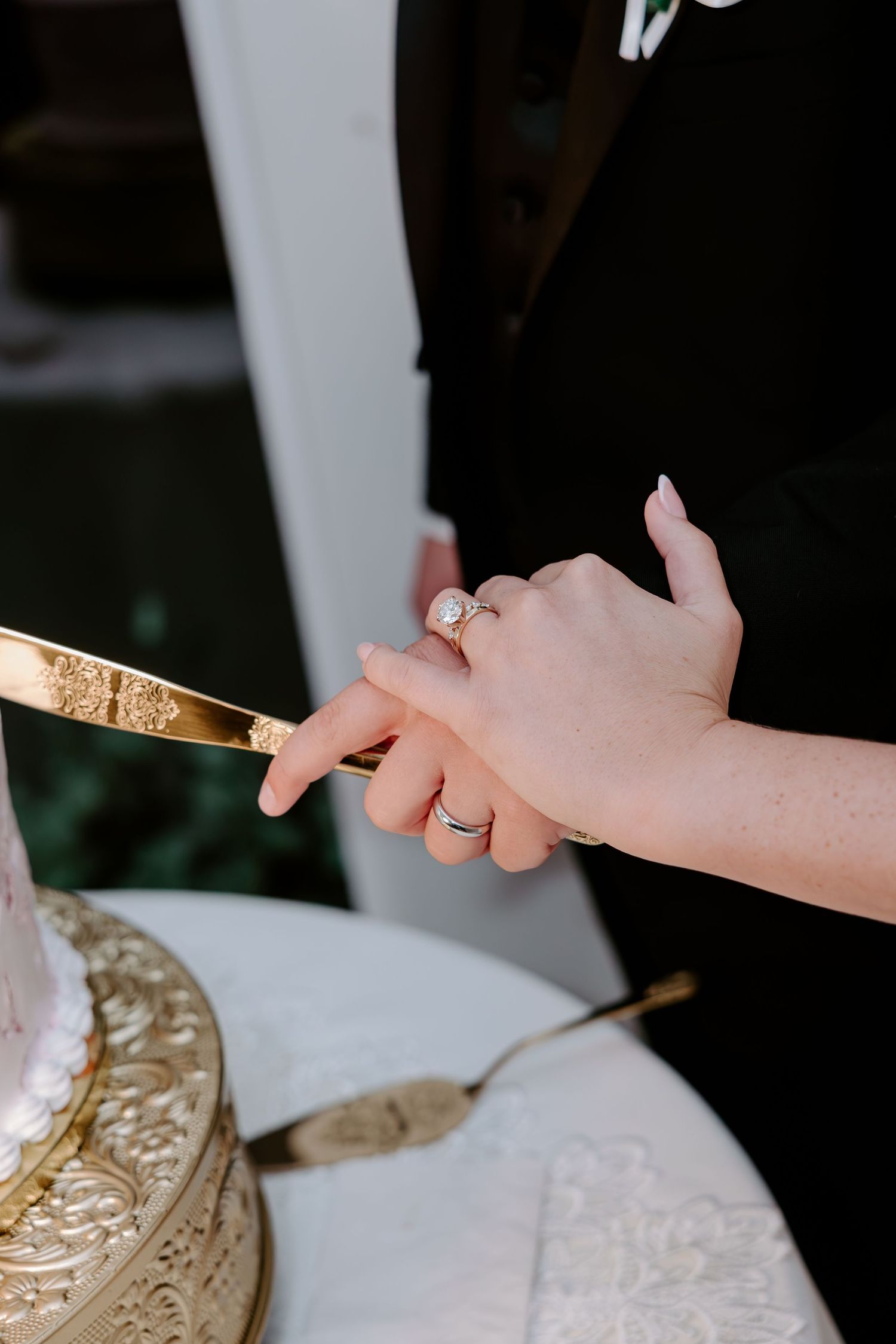 Newlyweds' hands with wedding rings hold a gold cake knife together to cut a tiered cake on an ornate gold stand.