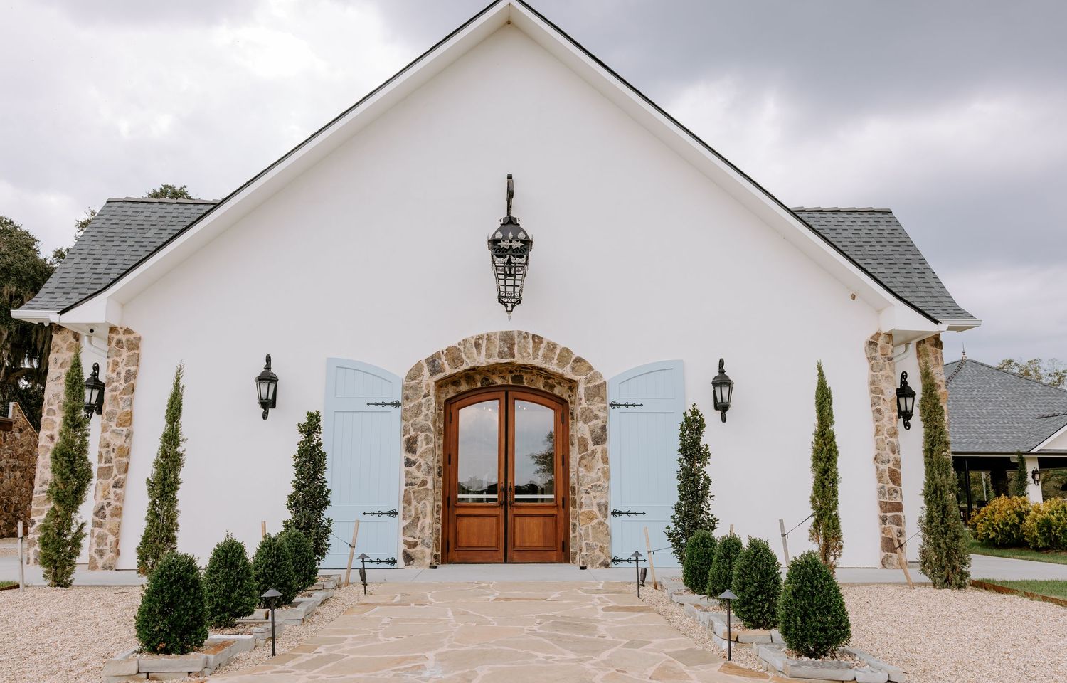 White chapel-style wedding venue with arched wooden double doors, stone accents, light blue shutters, and manicured evergreen shrubs.
