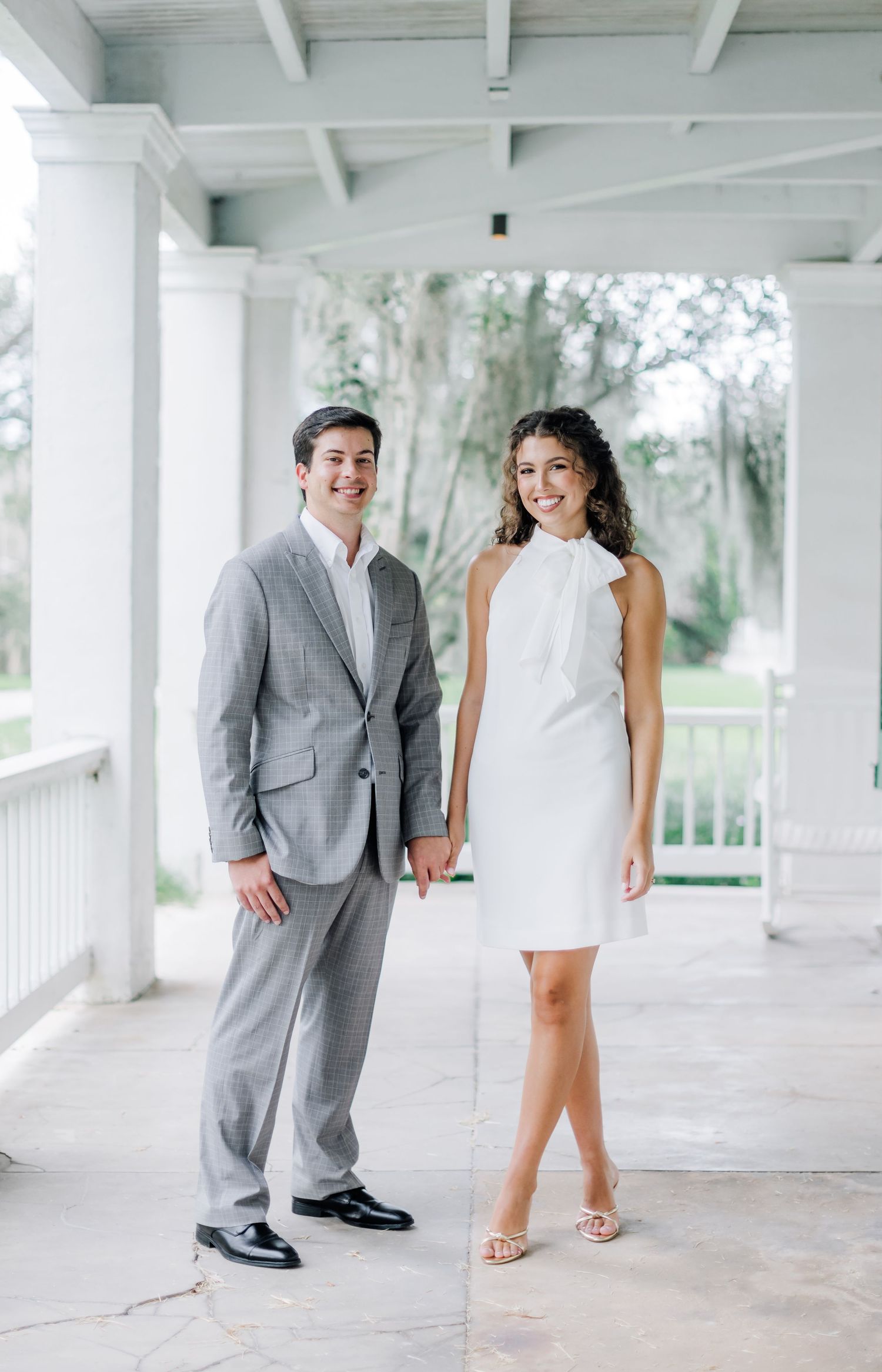 Couple in formal attire poses together on a white columned porch with trees in background.