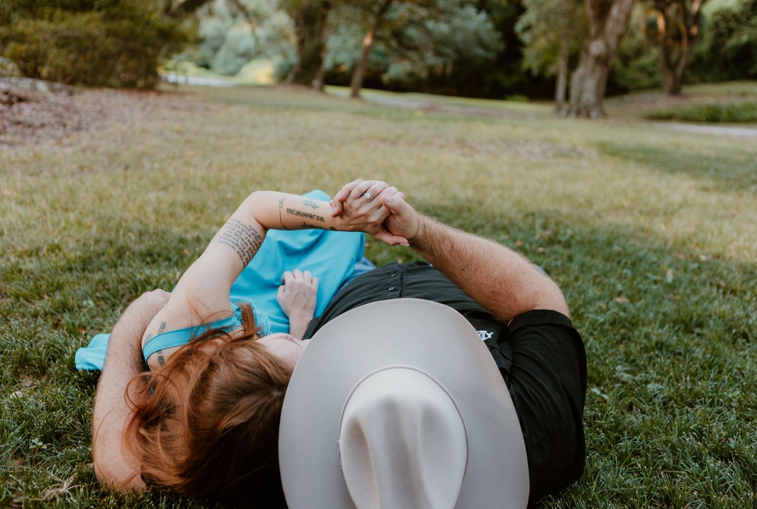 Person lying on grass wearing a white hat and turquoise shirt while looking at phone on a sunny day.