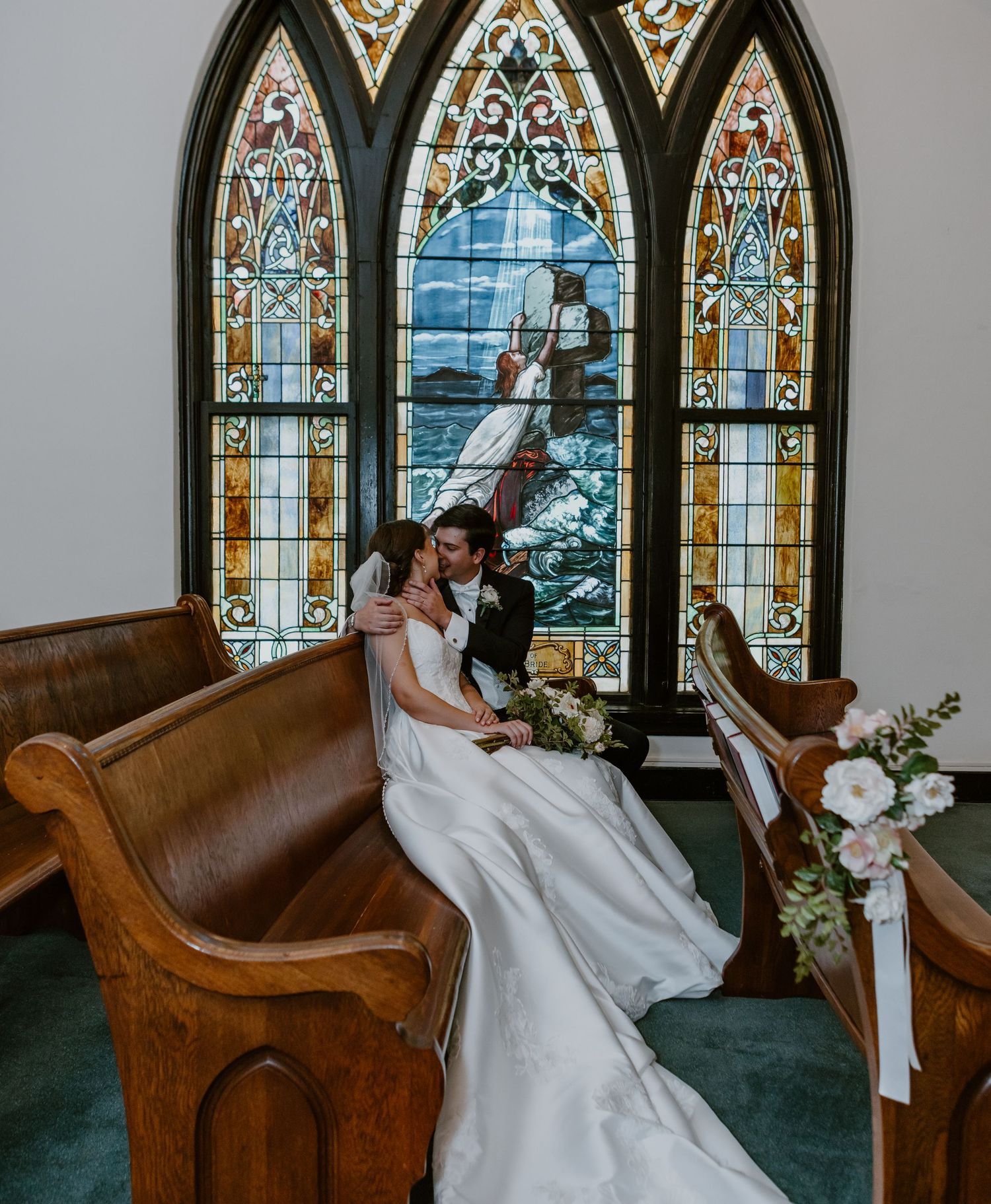 Bride and groom share an intimate moment on wooden church pews before a stunning stained glass window.