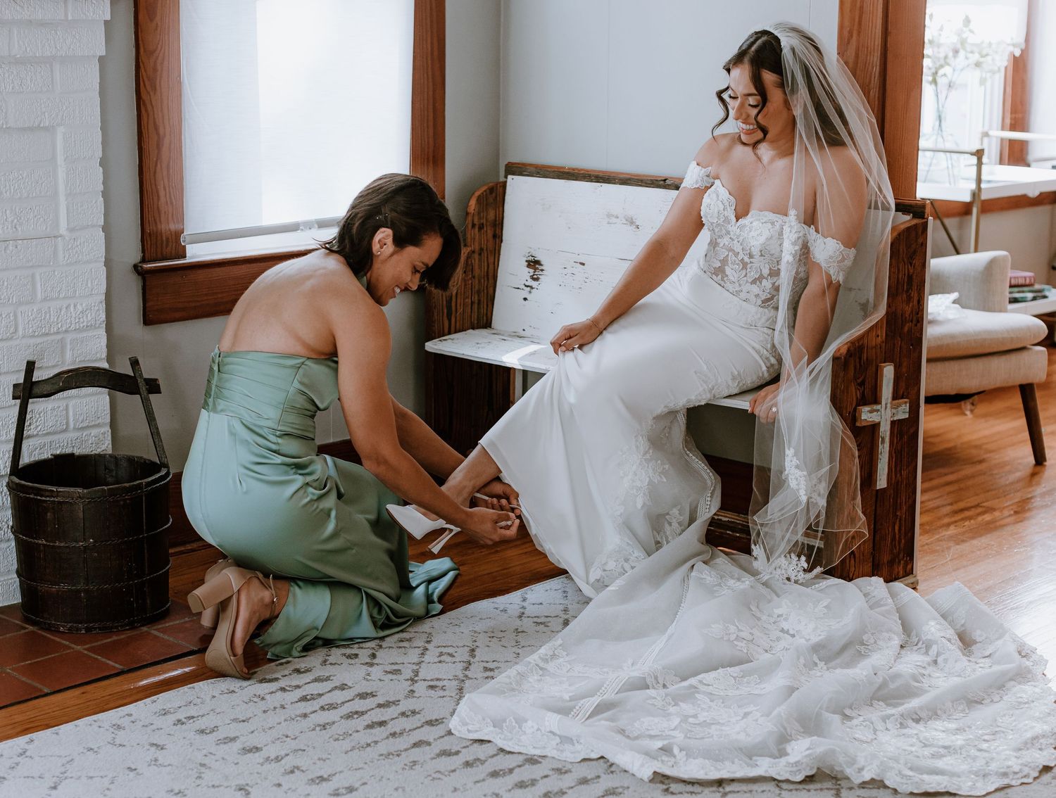 Bridesmaid in sage green dress helps bride in white gown with shoes while seated on bench with veil.
