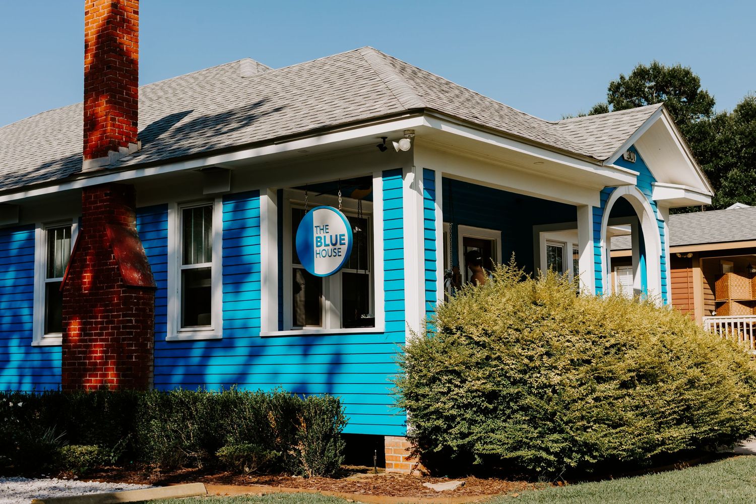 Bright blue cottage-style building with white trim, brick chimney, and lush green bushes on a sunny day.
