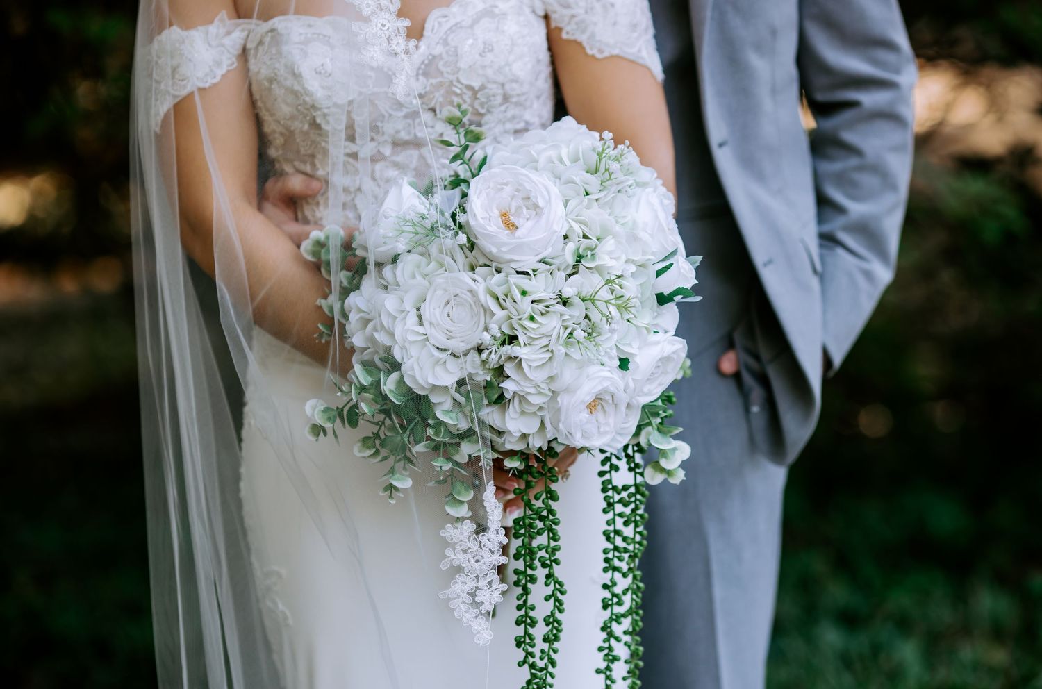 Bride holding a cascading white peony and greenery wedding bouquet beside a groom in a gray suit.