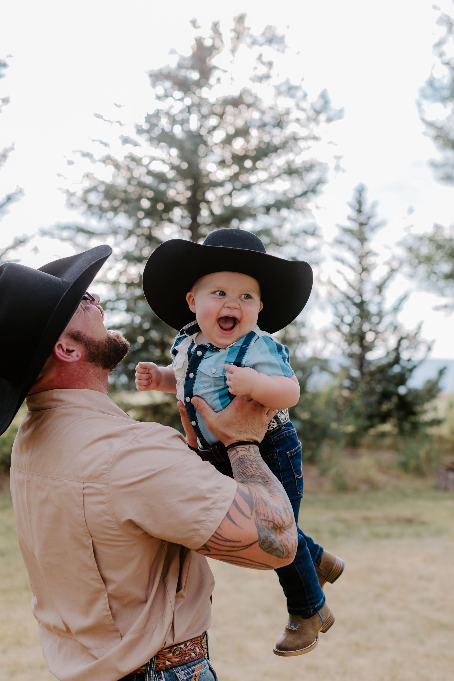 A tattooed man in a black cowboy hat lifts a laughing baby wearing a matching hat outdoors near pine trees.