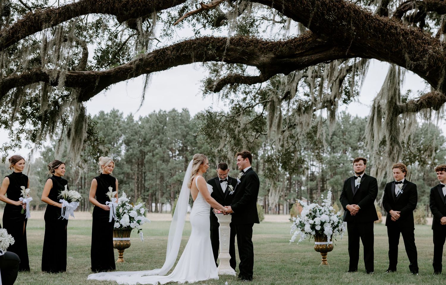 Bride and groom exchange vows under a mossy oak tree at an outdoor wedding ceremony with bridesmaids and groomsmen standing alongside.