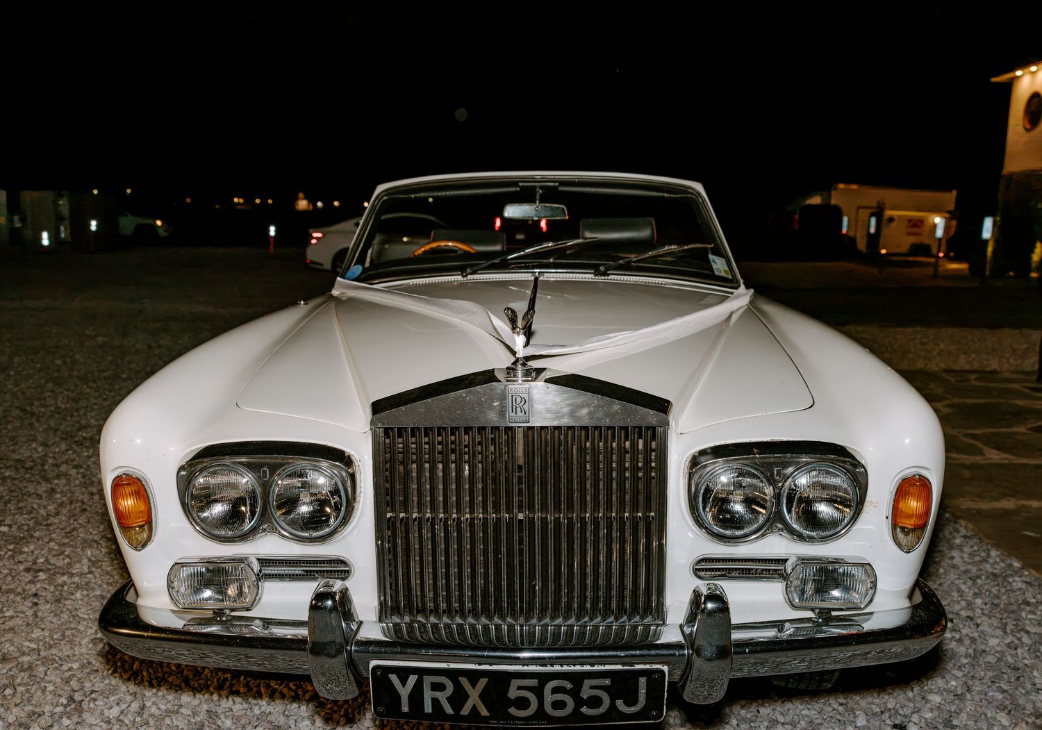 Front view of a white vintage Rolls-Royce convertible with chrome grille, round headlights and UK license plate parked outdoors at night.