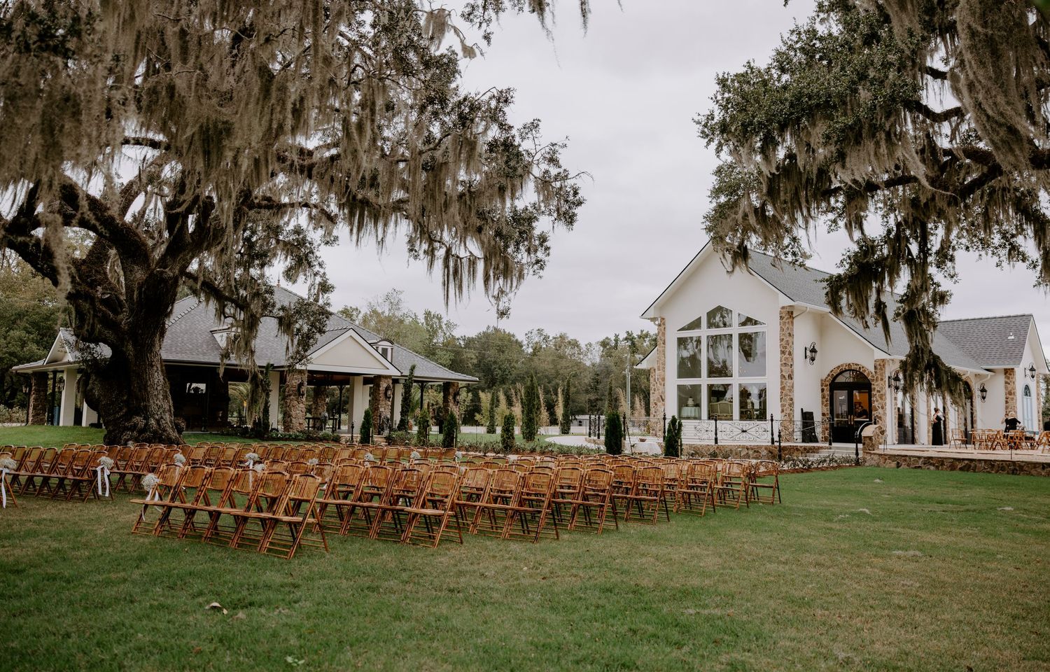 Outdoor wedding ceremony setup with rows of wooden chairs on a lawn beneath live oak trees draped in Spanish moss near a white chapel venue.